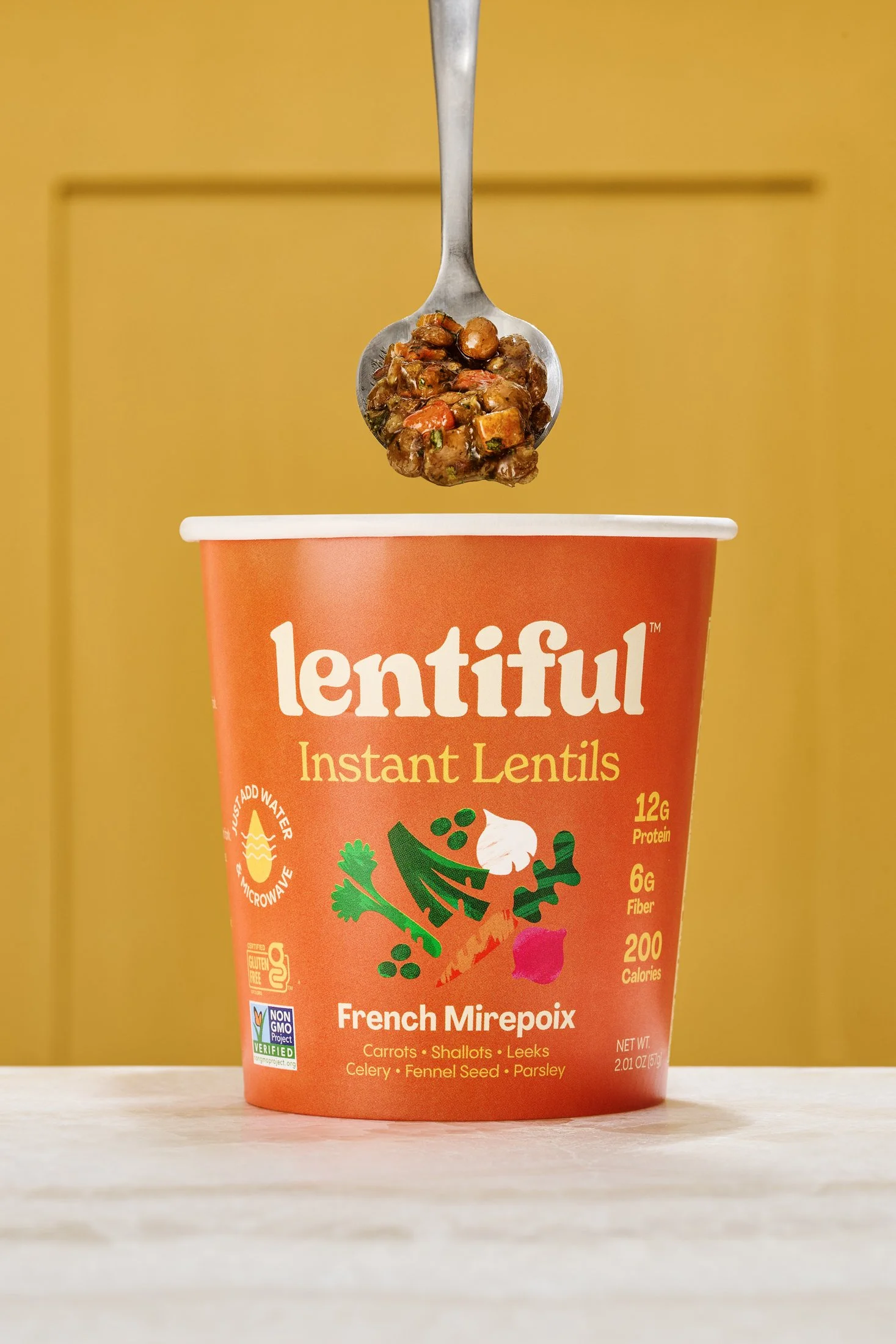 A cup of lentil soup with a spoonful being lifted above the cup, set against a yellow background. Product CPG food photography by Nutmeg Imageworks.