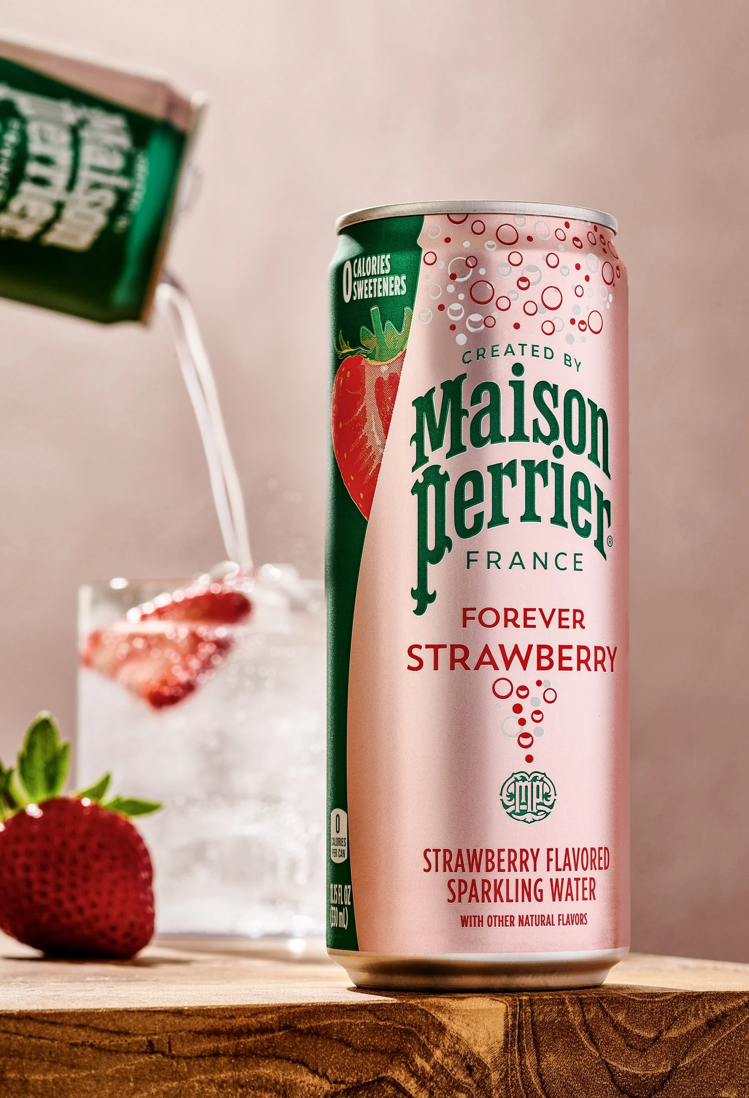 A pink can of Maison Quertier Forever Strawberry sparkling water with bubbles and strawberry graphics in foreground, and a blurred glass of strawberry soda with strawberries and mint in the background.