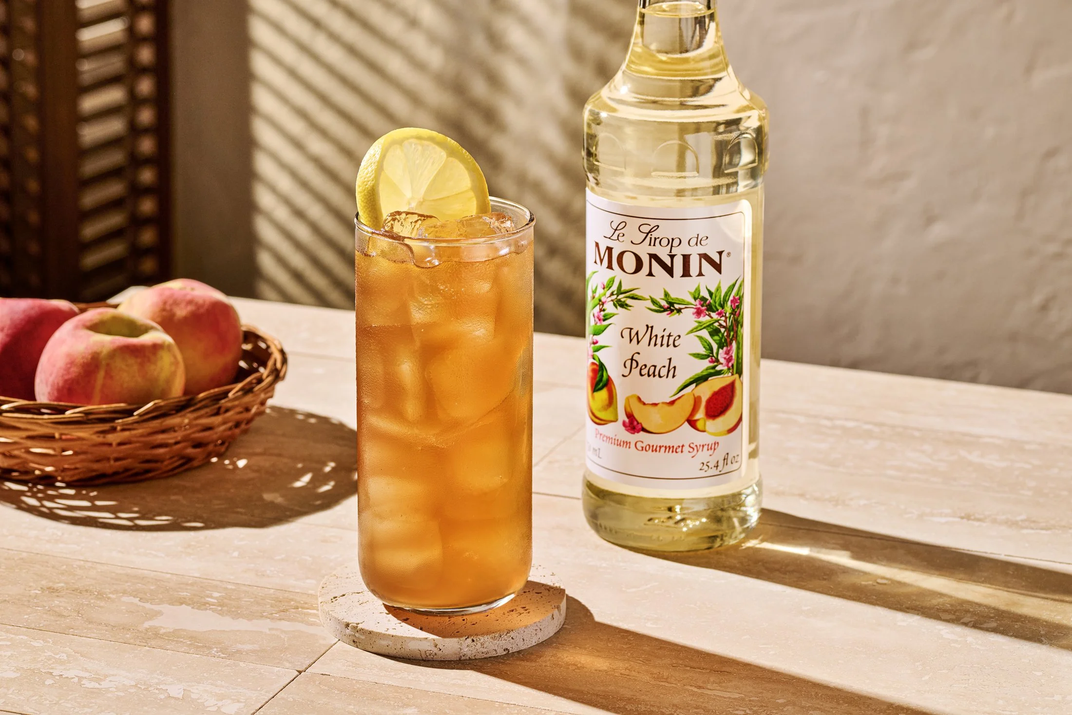 Beverage photography for Monin of a glass of iced tea garnished with a lemon slice, a bottle of Monin White Peach syrup, and a basket of peaches on a wooden surface with warm lighting.