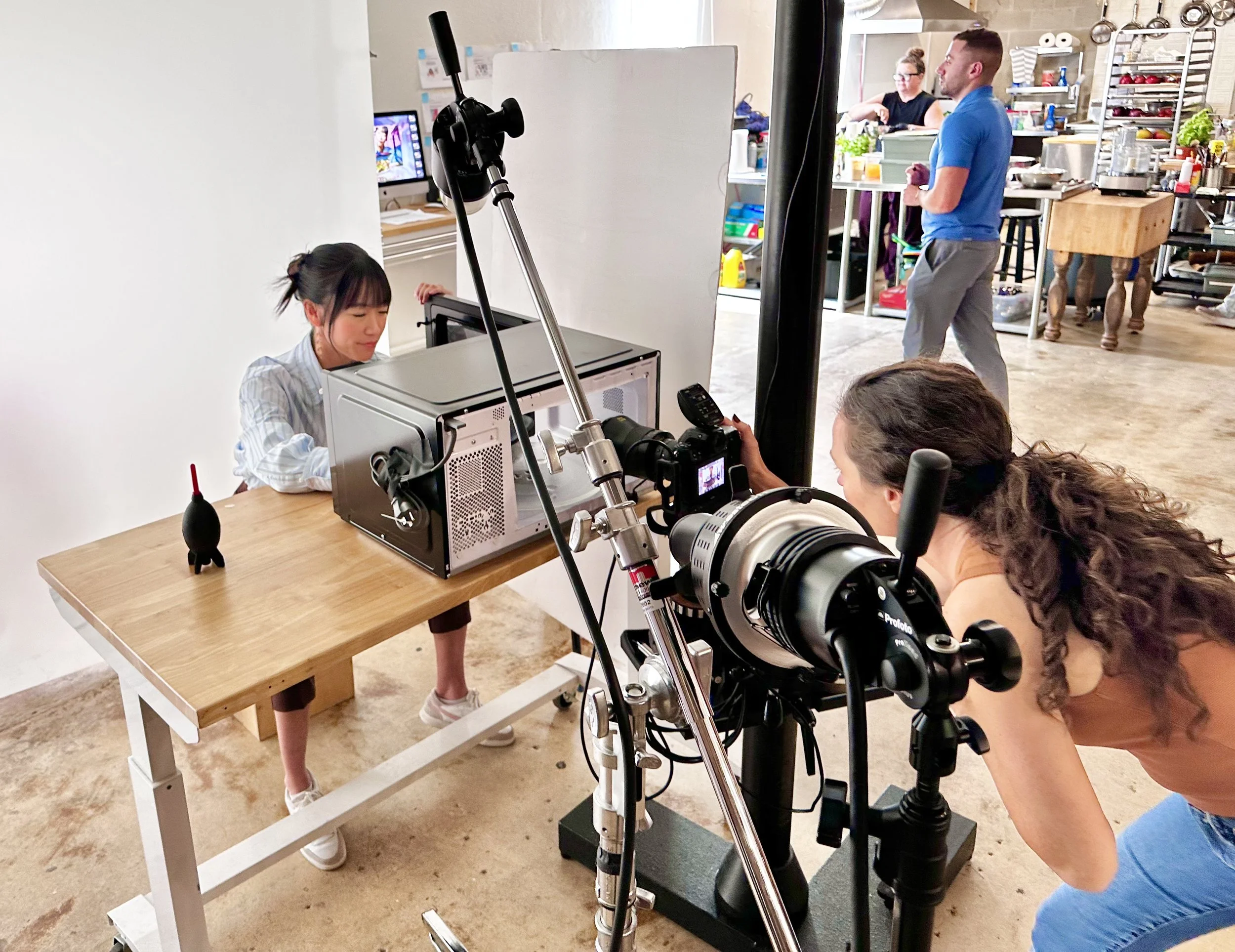Behind-the-scenes photo of a commercial food photoshoot, with a woman posing in front of a white backdrop, a camera on a tripod capturing her image, and crew members in the background in a kitchen setting.