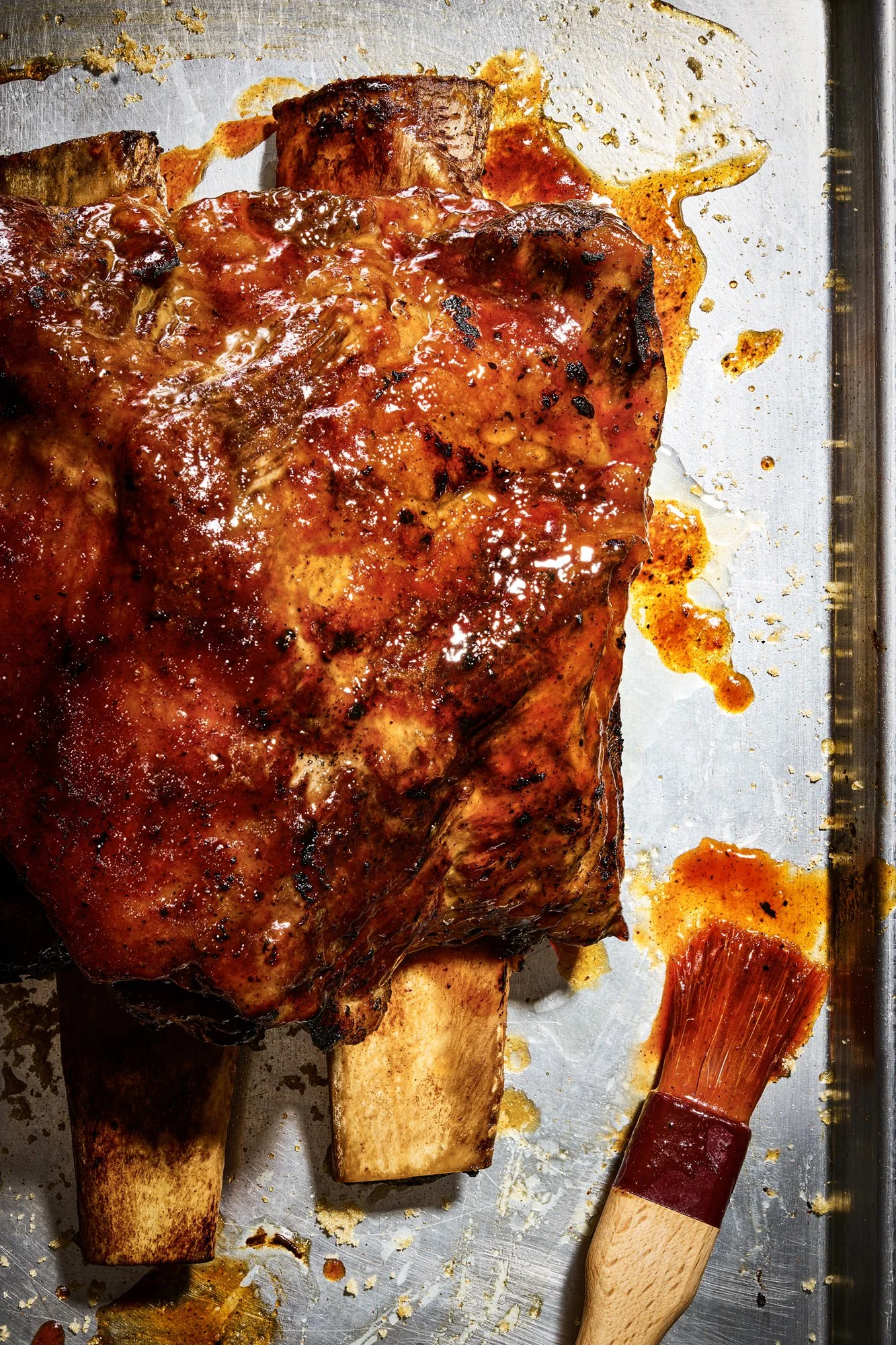 Grilled meat, possibly ribs, on a metal tray with a meat bushing nearby, sauce and juices spilled on the tray.