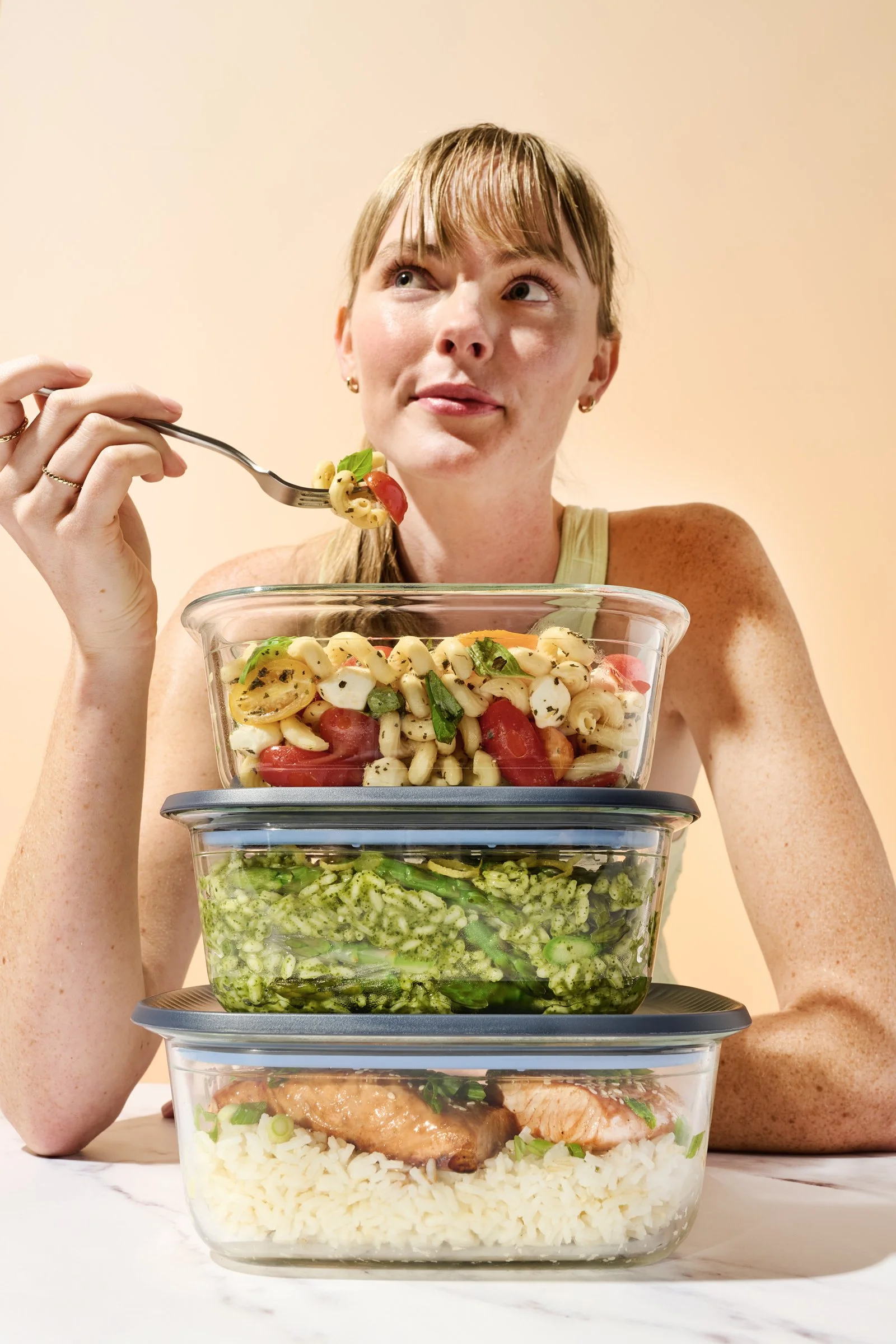 A woman sitting at a table with three stacked glass meal containers filled with salad and cooked chicken, holding a fork with pasta salad, looking thoughtfully.