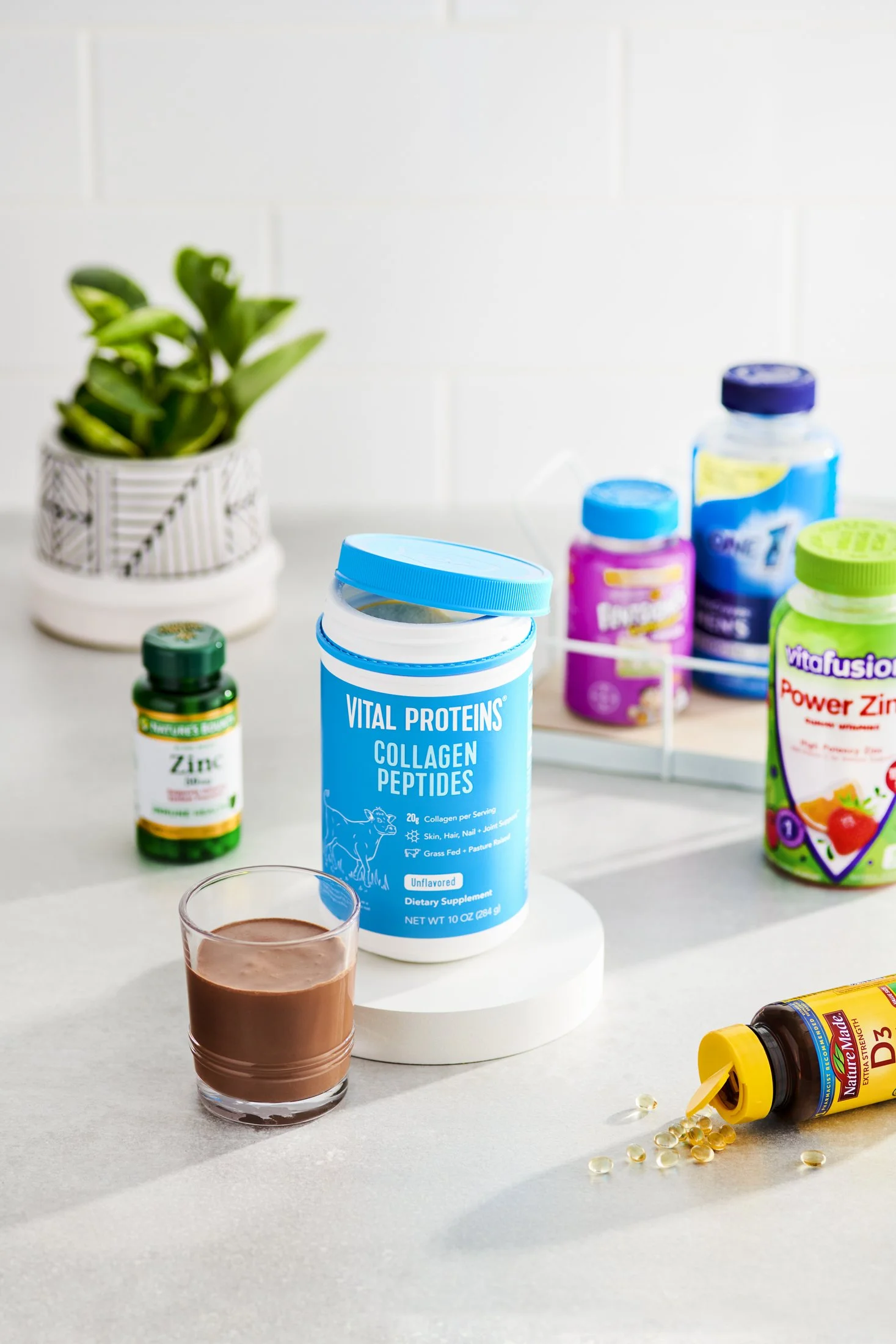 A glass of chocolate drink in the foreground with various supplement bottles on a white surface, a potted plant in a black and white striped pot in the background, and a white brick wall behind.