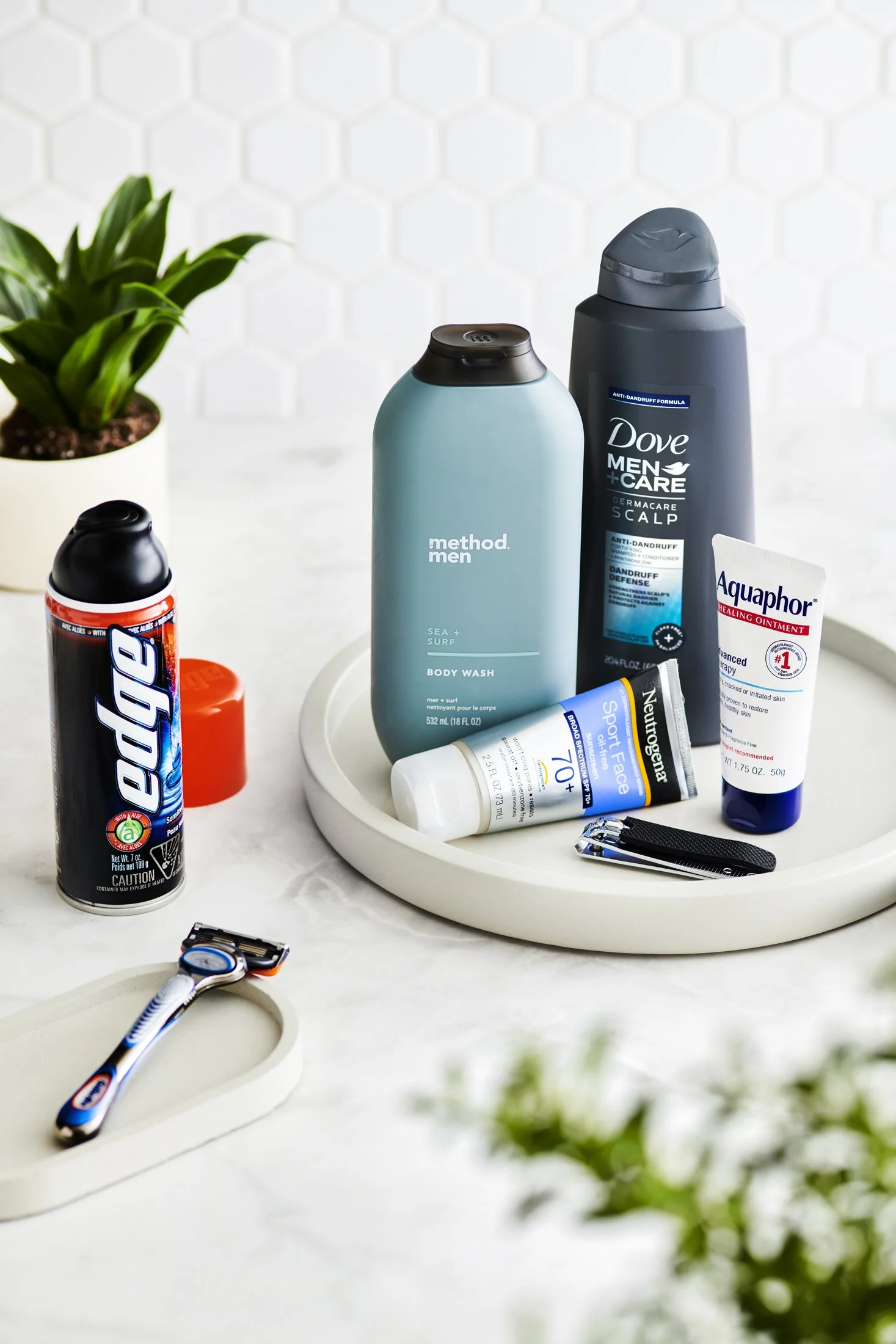 A bathroom countertop with various personal care items, including deodorant, face wash, scalp treatment, toothpaste, a razor, and a small potted green plant in the background.