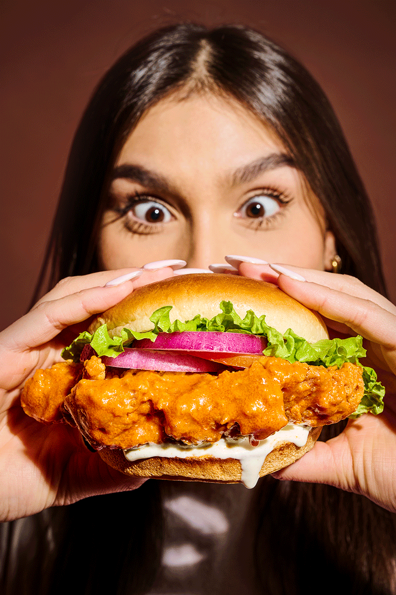A young woman with long dark brown hair and wide eyes holding a large fried chicken sandwich with lettuce, pickles, onions, and sauce at the top.
