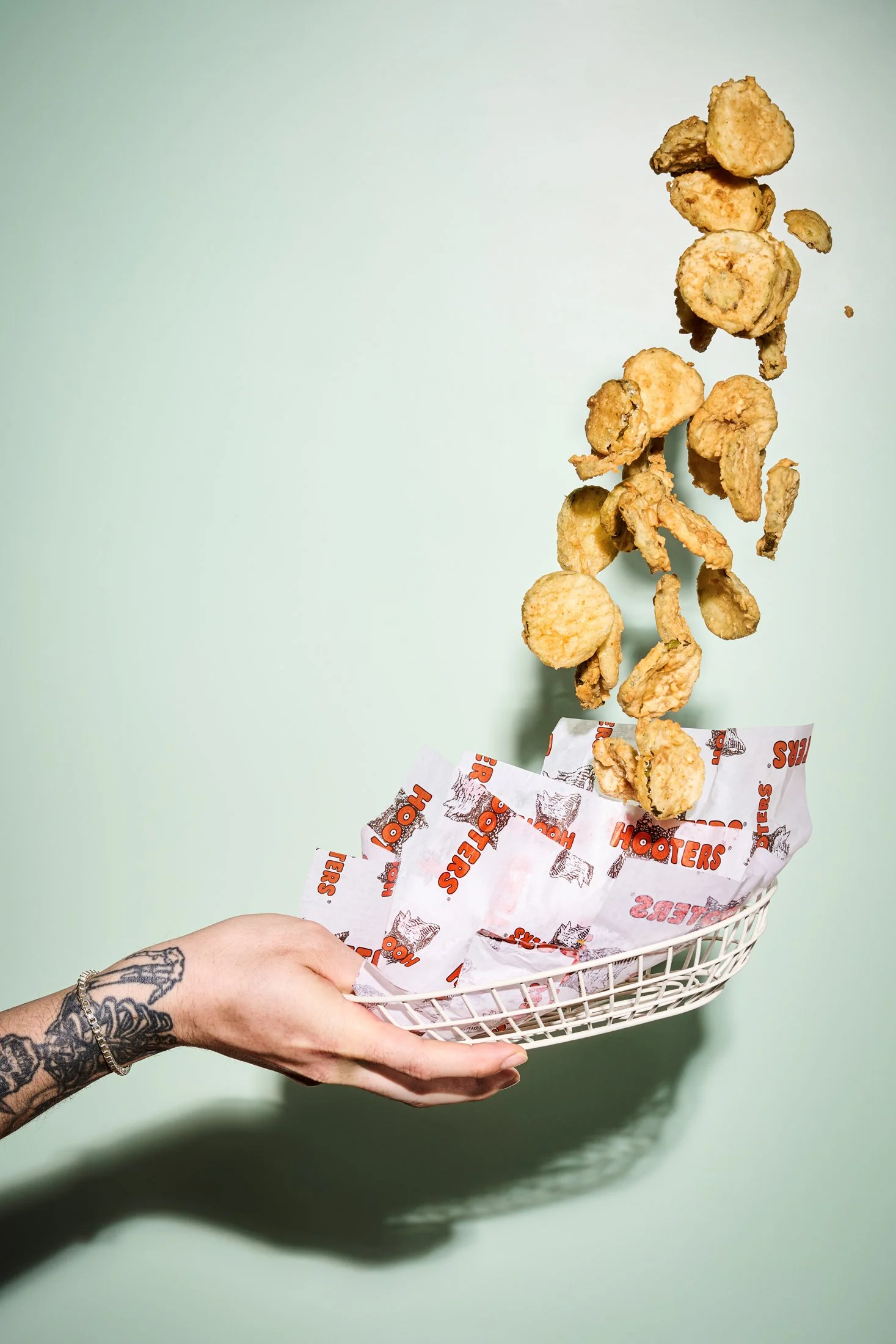 Fried chicken pieces falling into a basket lined with Hooters-branded paper, held by a person with a tattooed hand and bracelet, against a mint green background.