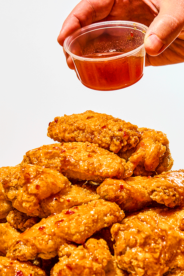 Close-up of a pile of crispy fried chicken pieces with a hand holding a small clear container of hot sauce above them.