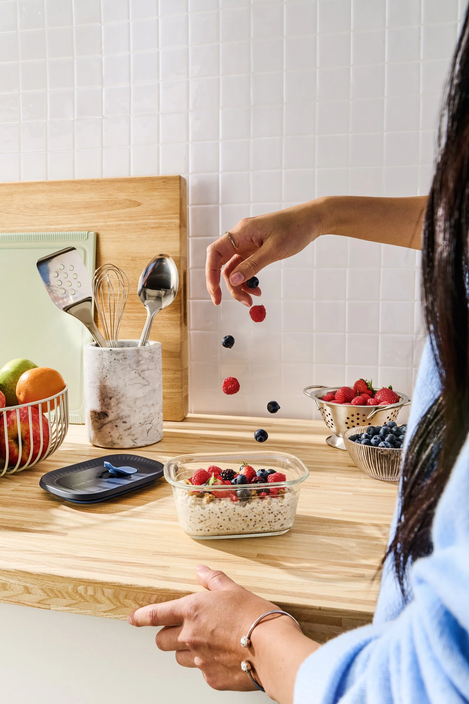 Person mid-air dropping mixed berries into a glass container of oatmeal topped with berries on a kitchen counter.