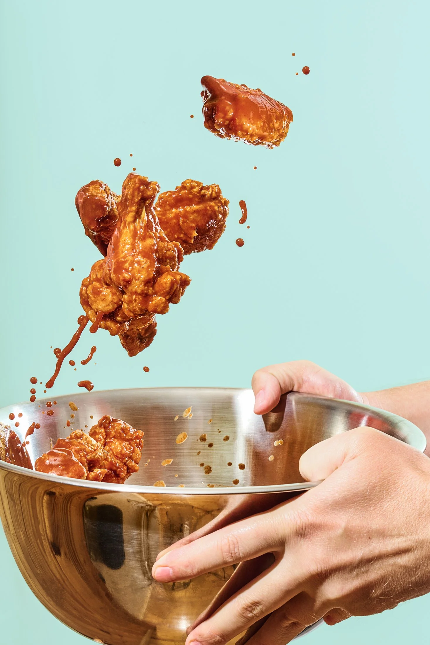 Fried chicken pieces tossed in sauce being dropped into a metal bowl, against a light blue background.
