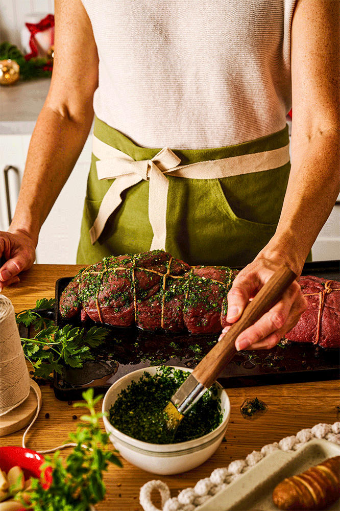 Person preparing a seasoned beef roast tied with kitchen twine, brushing herbs from a bowl, on a wooden kitchen table.