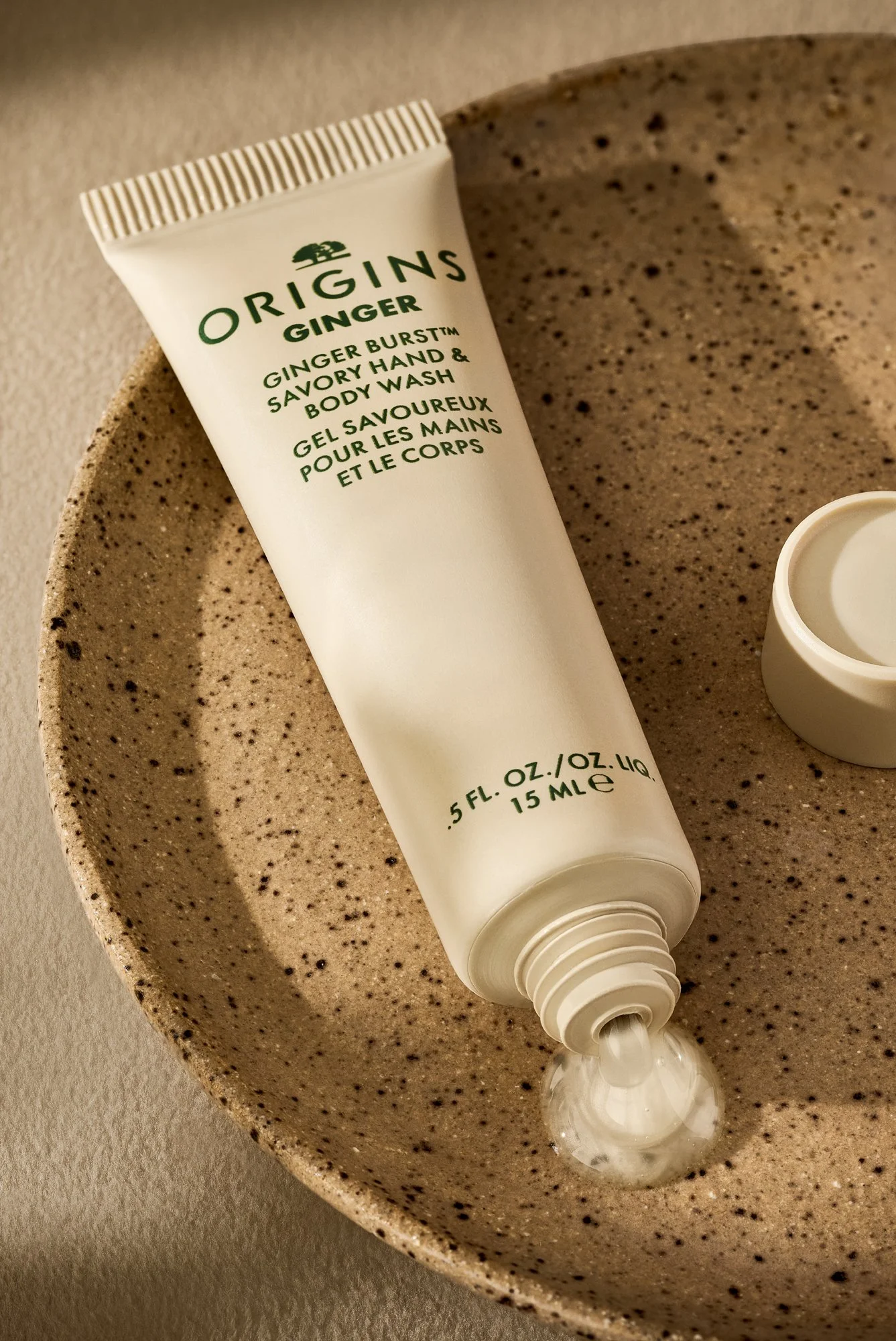 A tube of Origins body wash with some of the product coming out of the tube, resting on a speckled beige ceramic bowl. Product photography by Nutmeg Imageworks.