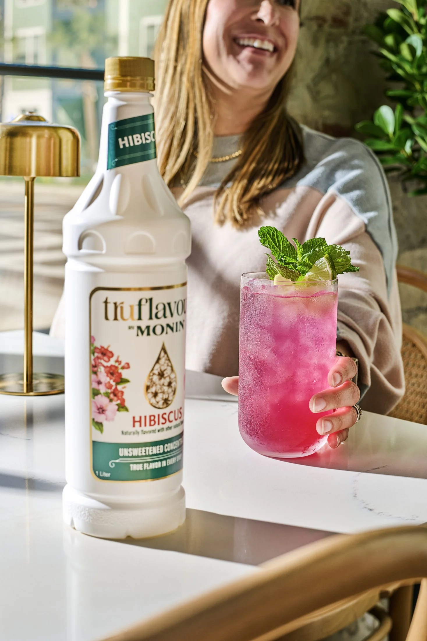 A woman sitting at a table holding a pink hibiscus flavored drink garnished with fresh mint and lime, with a bottle of hibiscus flavoring and a gold lamp in the foreground. Advertising lifestyle photography on location.