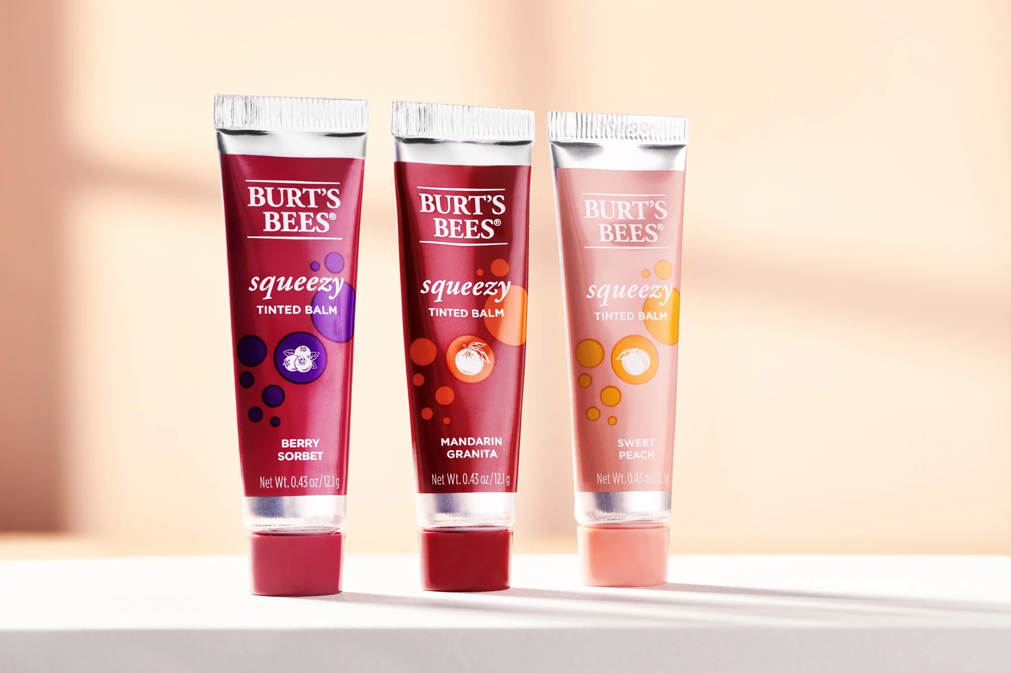 Three tubes of Burt's Bees tinted lip balm standing upright on a white shelf against a soft pink background. Beauty product photography by Nutmeg Imageworks.