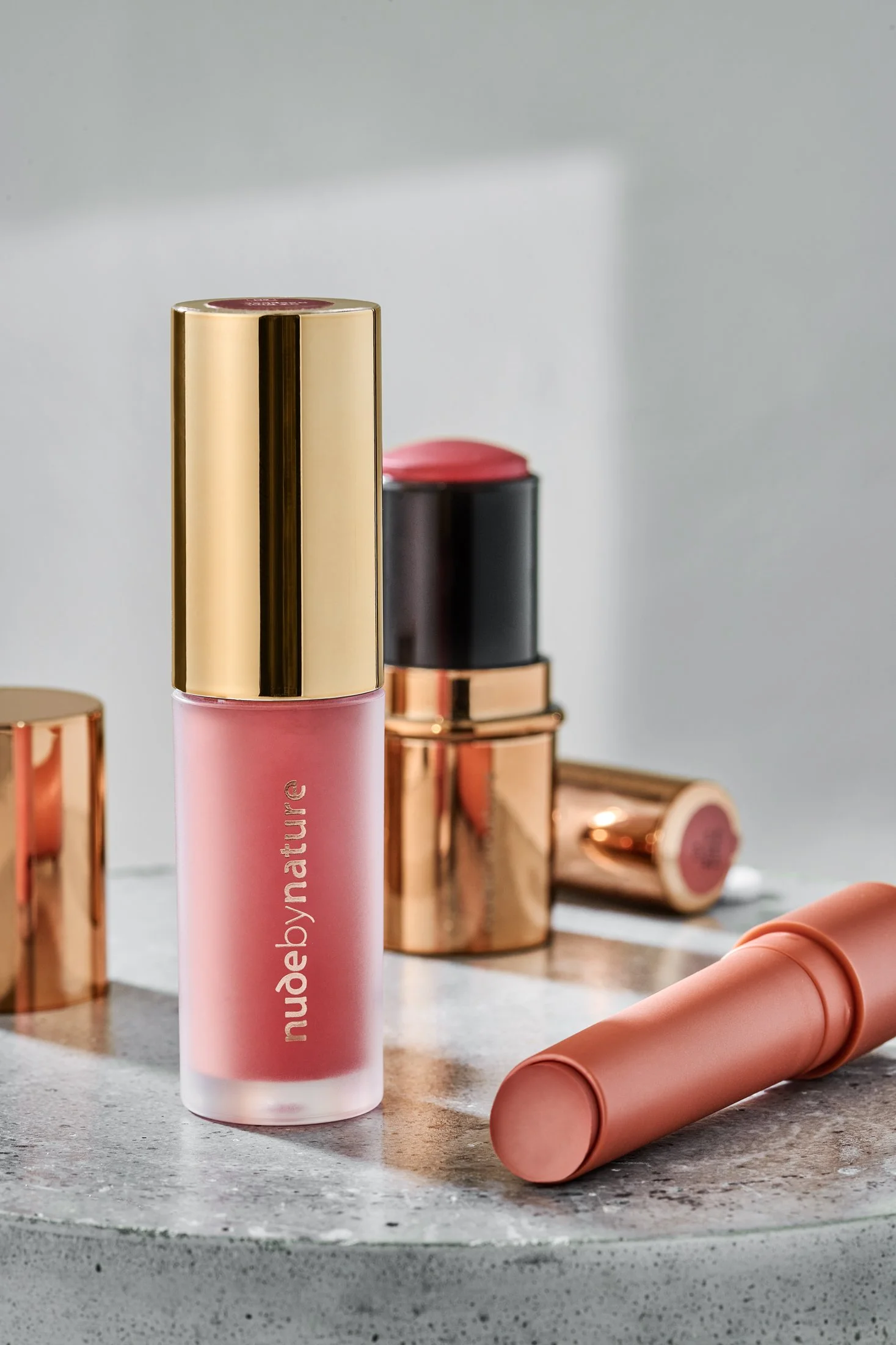 A pink cylindrical skincare product with a shiny gold cap labeled 'nude by nature' on a textured gray surface, surrounded by various lipstick or lip balm tubes in shades of gold and pink, some lying on their sides and some standing upright.