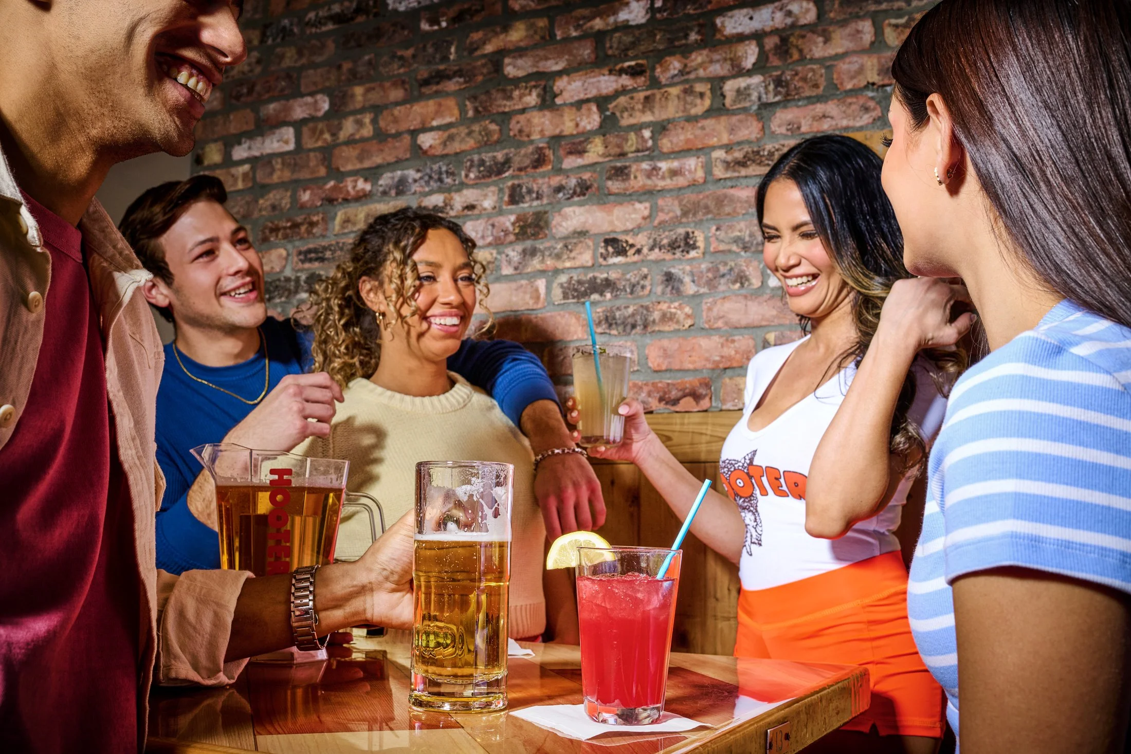 A group of five friends gathered around a bar table bar at a restaurant, smiling and enjoying drinks, with a brick wall in the background.