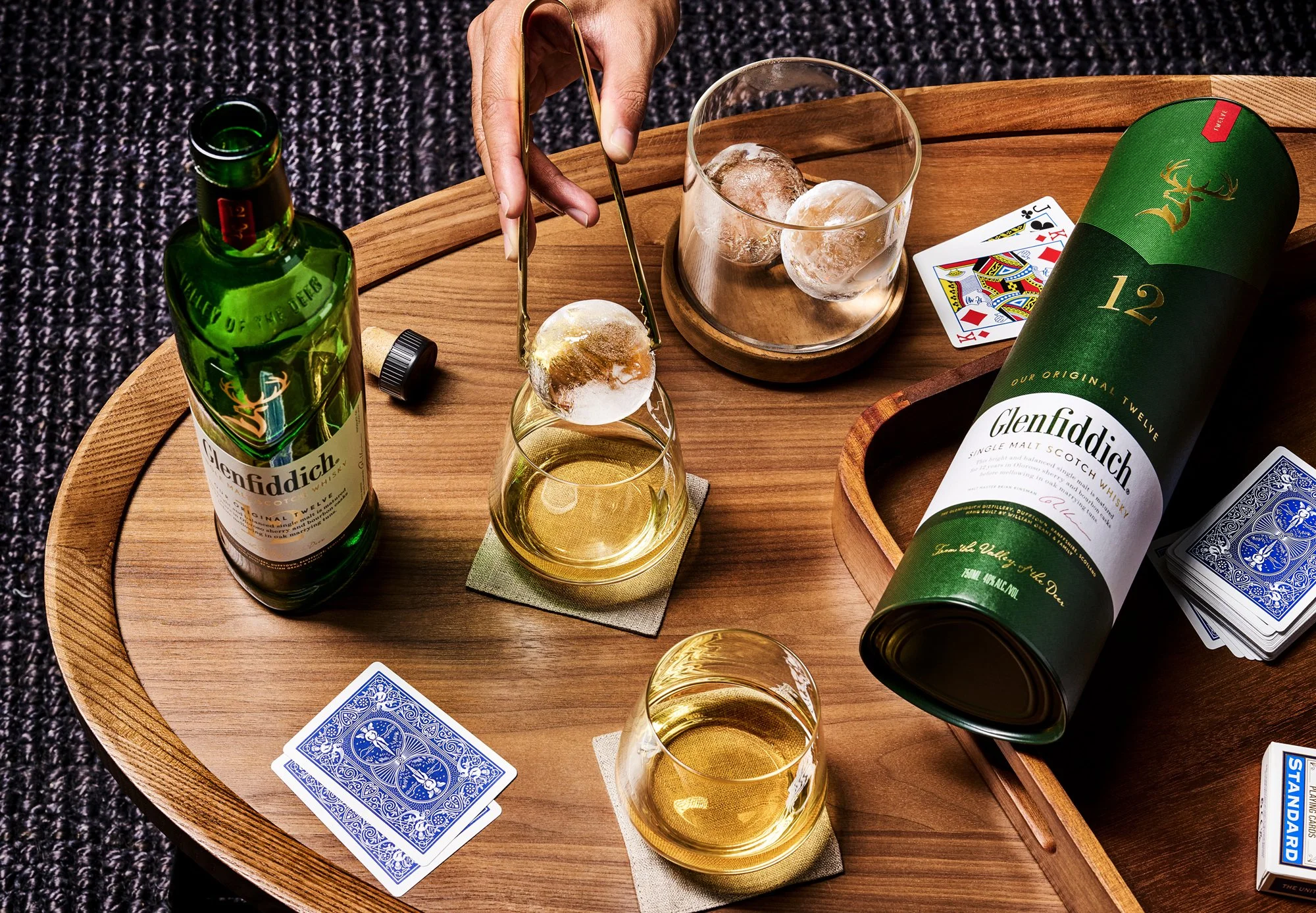 A wooden tray with a bottle of Glenfiddich single malt Scotch whisky, two glasses of whisky, ice cubes, playing cards, and a hand using tongs to place ice into a glass. Product beverage photography by Nutmeg Imageworks.