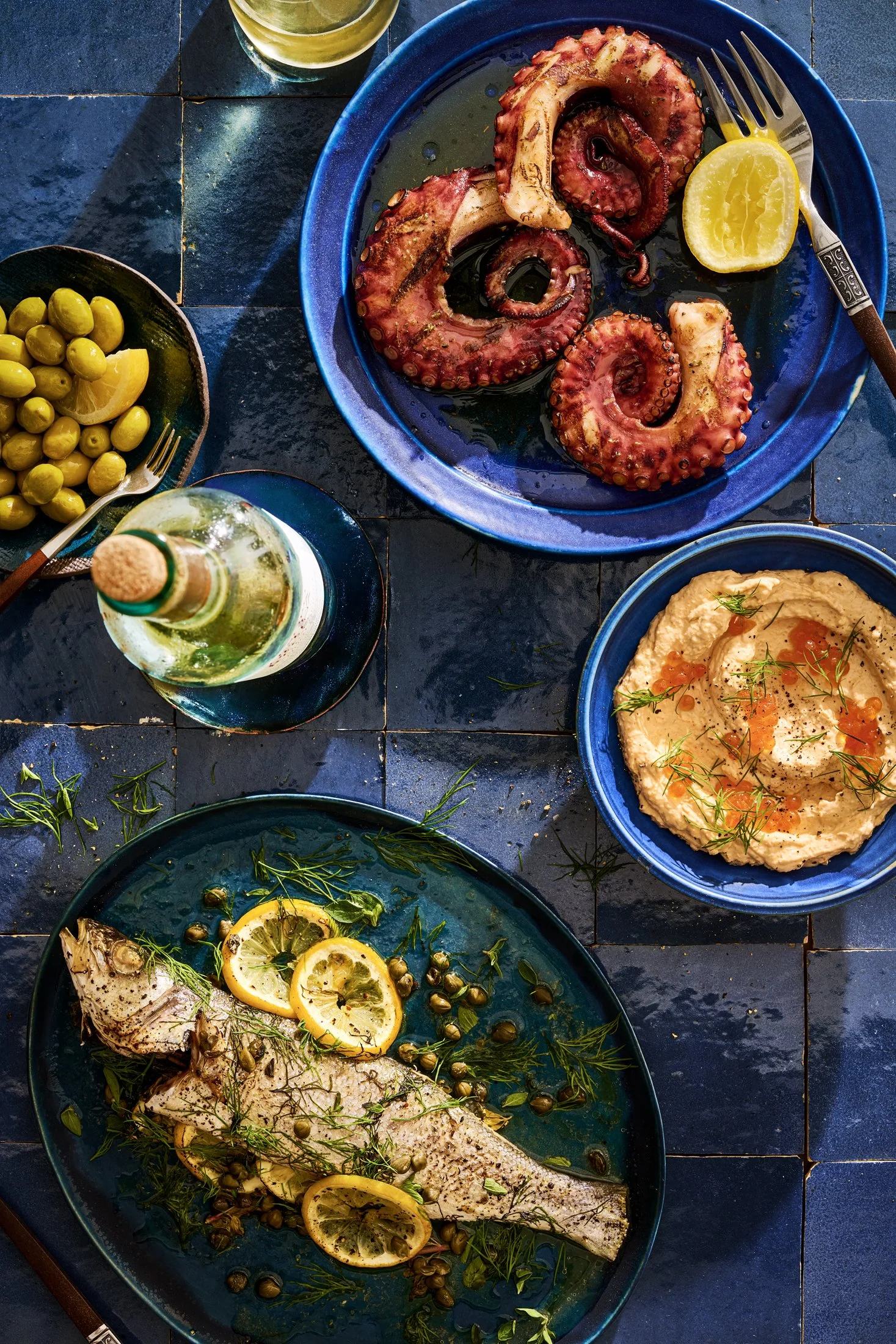 A Mediterranean seafood meal with grilled octopus on a blue plate, a whole baked fish garnished with lemon slices and herbs, a bowl of hummus topped with salmon roe and dill, a small bowl of green olives with a lemon wedge, and a glass of white wine 