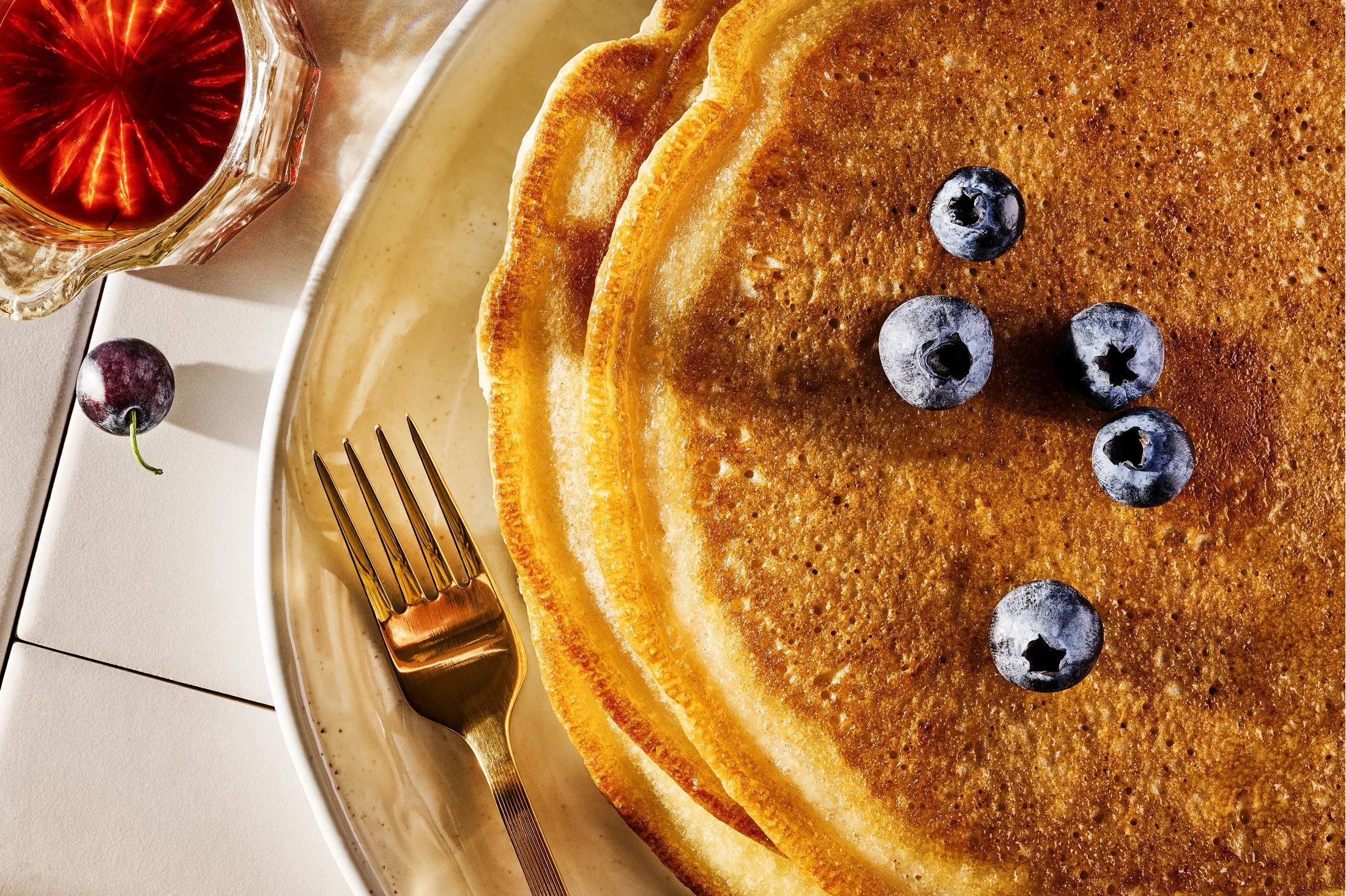 A stack of three golden brown pancakes topped with blueberries in a white bowl. To the side, there is a fork and a glass of red syrup or juice. The setting is on a light-colored surface.
