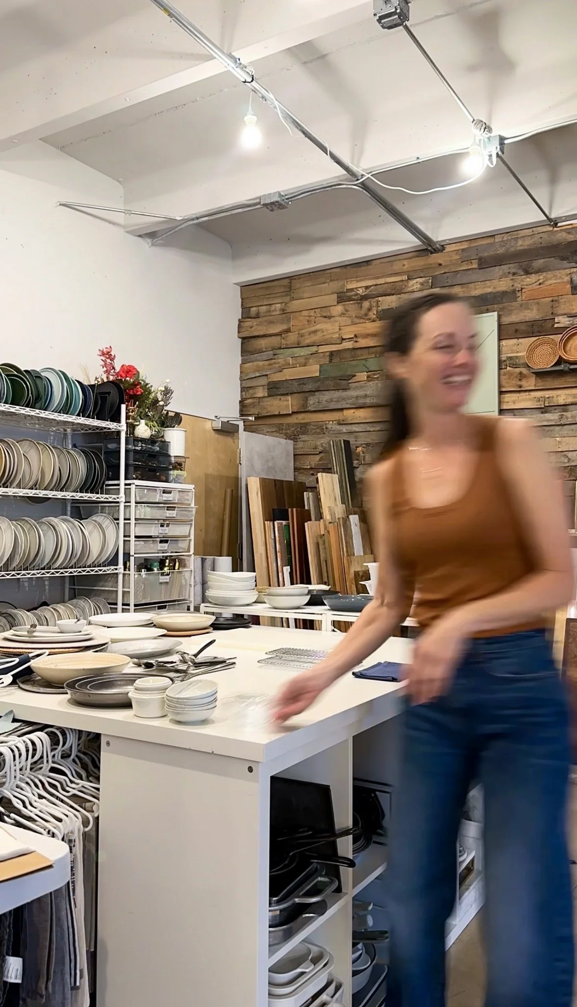 A woman smiling and reaching for white and gray ceramic dishes on a white kitchen island in a store, with shelves of plates and bowls, wood accents on the wall, and kitchenware underneath.