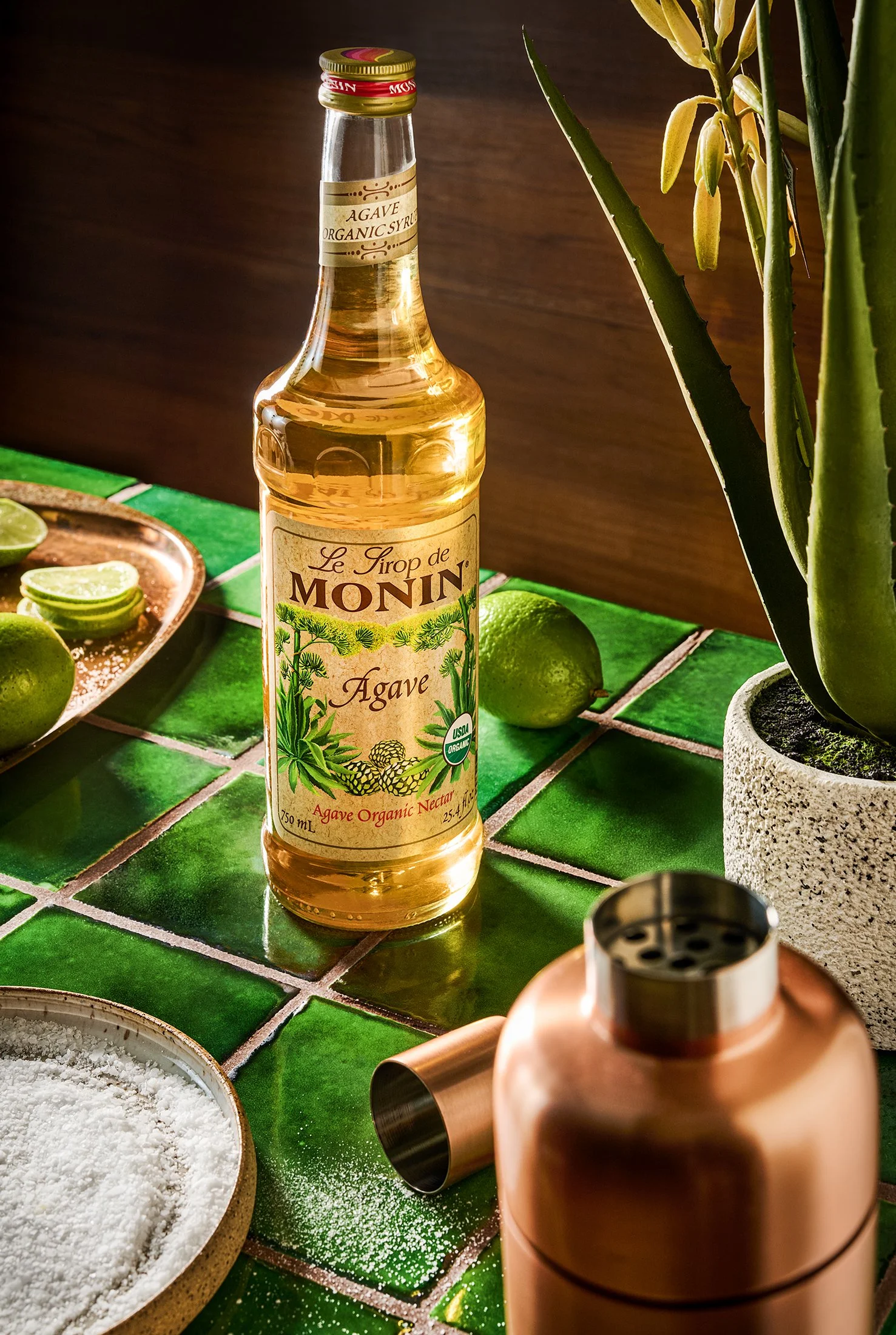 A bottle of agave organic syrup on a green tiled kitchen countertop with limes, a small bowl of salt, and a copper cocktail shaker.
