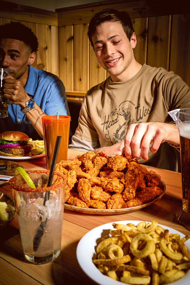 Two men at a table with fried chicken, French fries, a burger, and drinks, smiling and enjoying a meal. Advertising photography lifestyle.