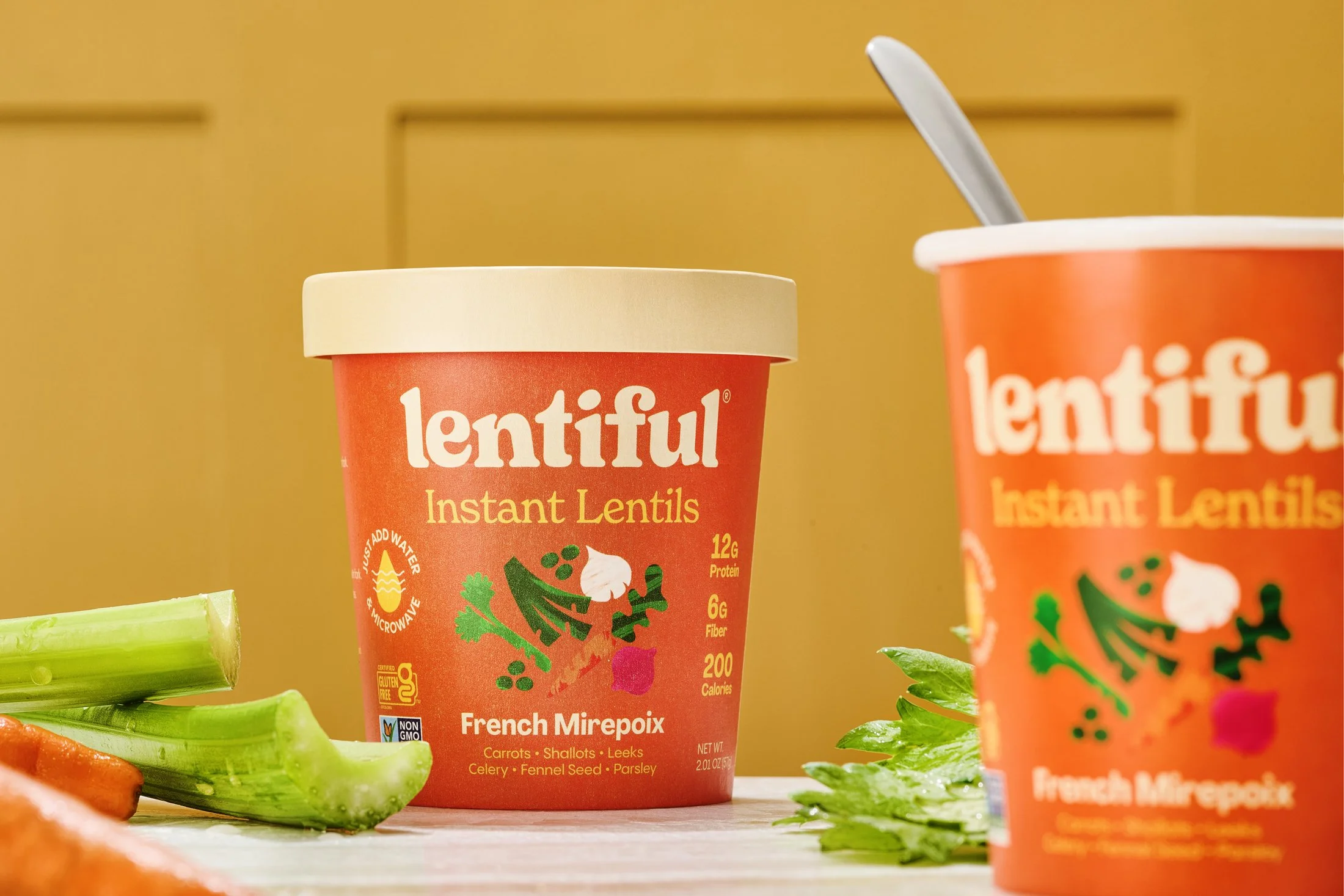 A cup of lentil soup with fresh vegetables around, set against a yellow background. Product CPG food photography by Nutmeg Imageworks.