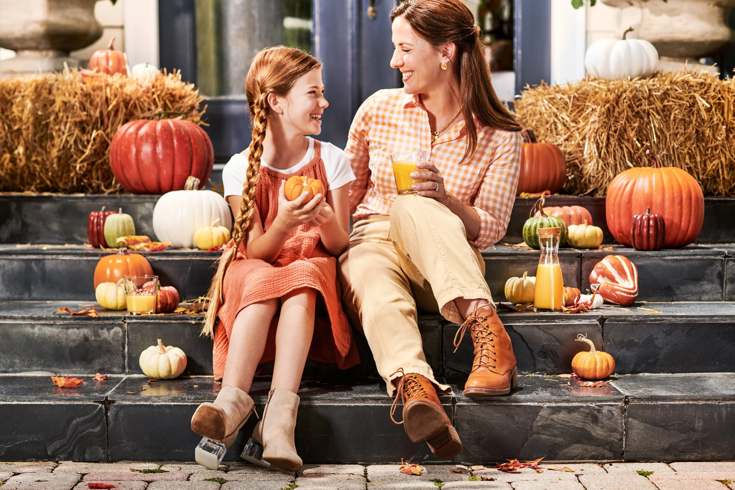 A woman and girl sitting on porch steps surrounded by pumpkins and fall decorations, sharing a moment and smiling at each other.
