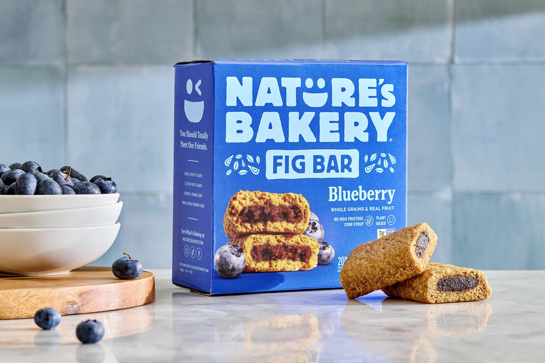 A box of Nature's Bakery blueberry fig bars with two bars placed in front. To the left, a bowl of fresh blueberries with some spilling onto a marble surface.