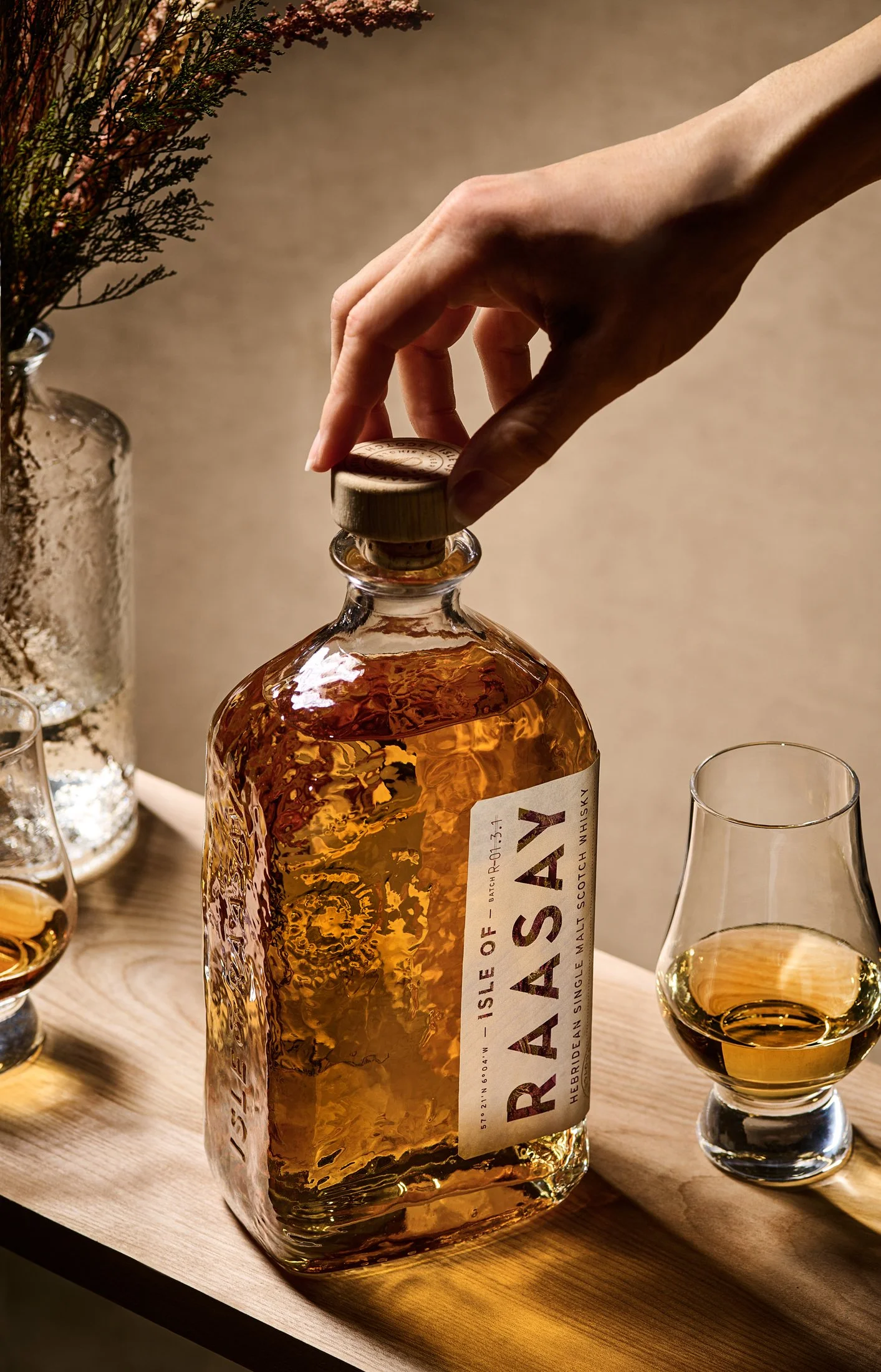 A hand opening a bottle of RAREST single malt Scotch whisky labeled 'Isle of Arran' on a wooden table with whiskey glasses and a vase with dried flowers.