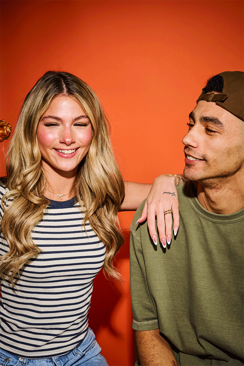 A young woman with long blonde hair smiling and wearing a striped shirt, standing with her arm around a young man wearing a green t-shirt and a backward cap against an orange background.
