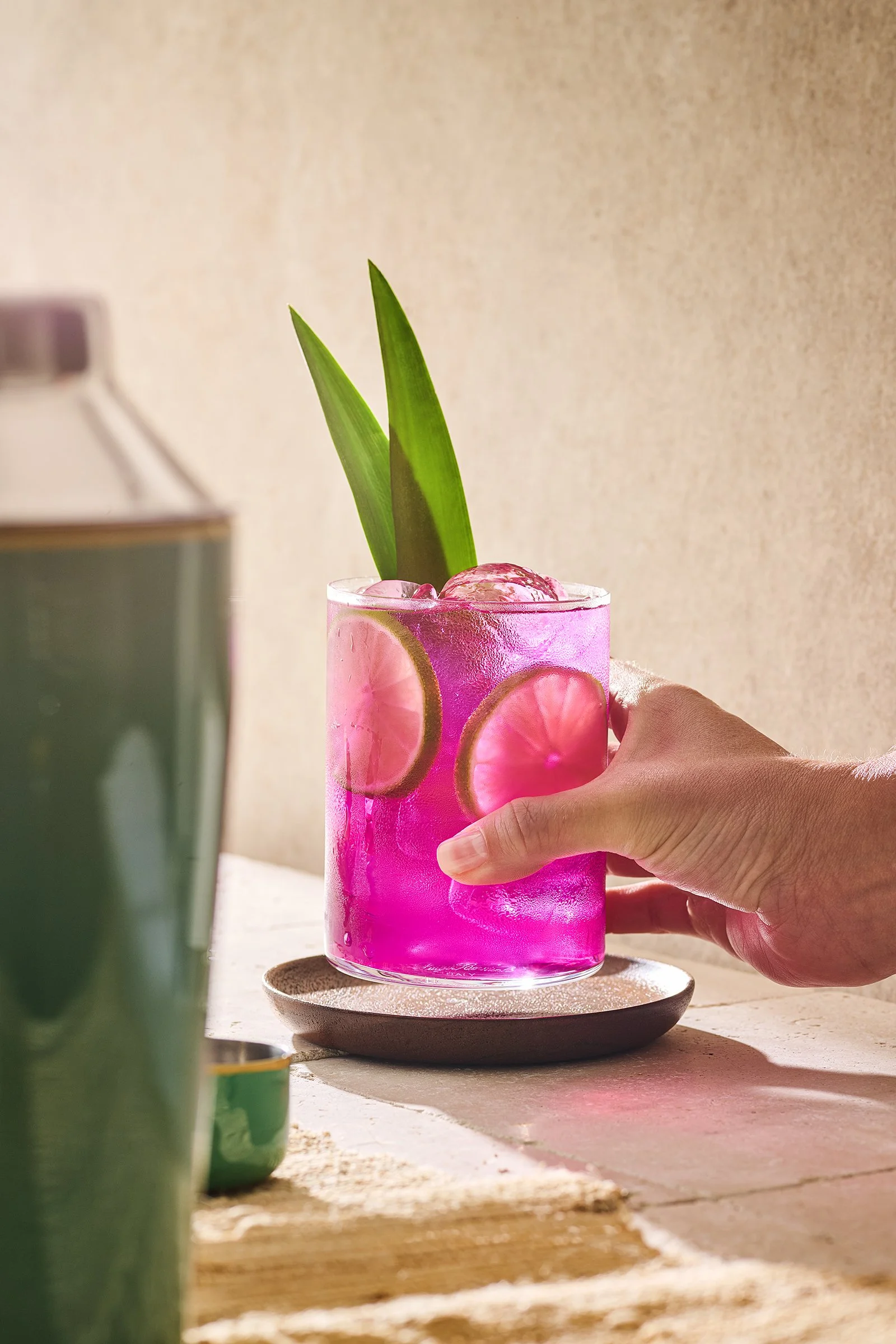 A hand holding a glass of bright pink drink with lemon and lime slices, ice, and two green leaves as garnish, on a round tray.