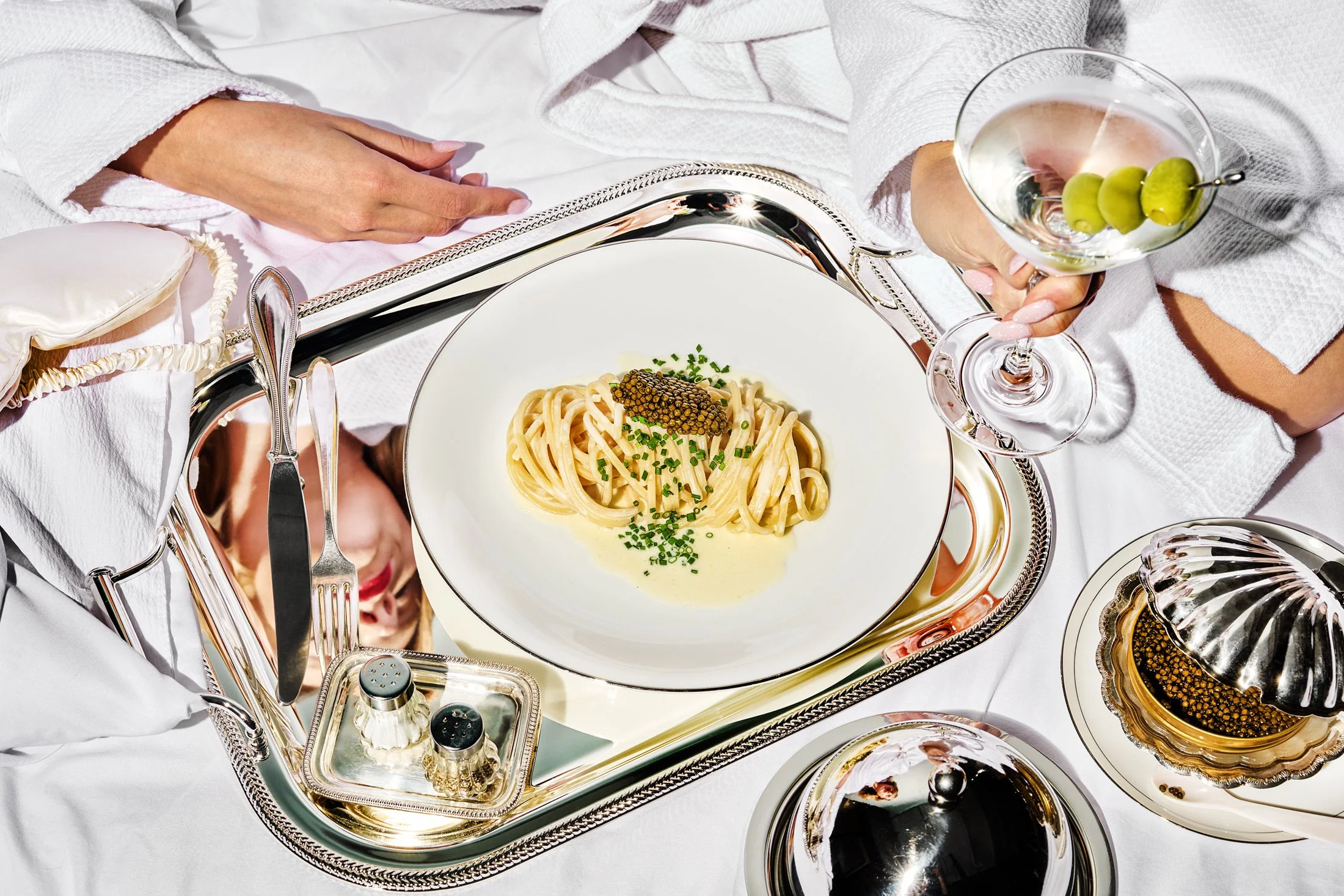 Elegant table setting with a white plate of pasta garnished with green herbs, surrounded by silverware, a martini glass with olives, and other silver serving dishes on a white tablecloth.