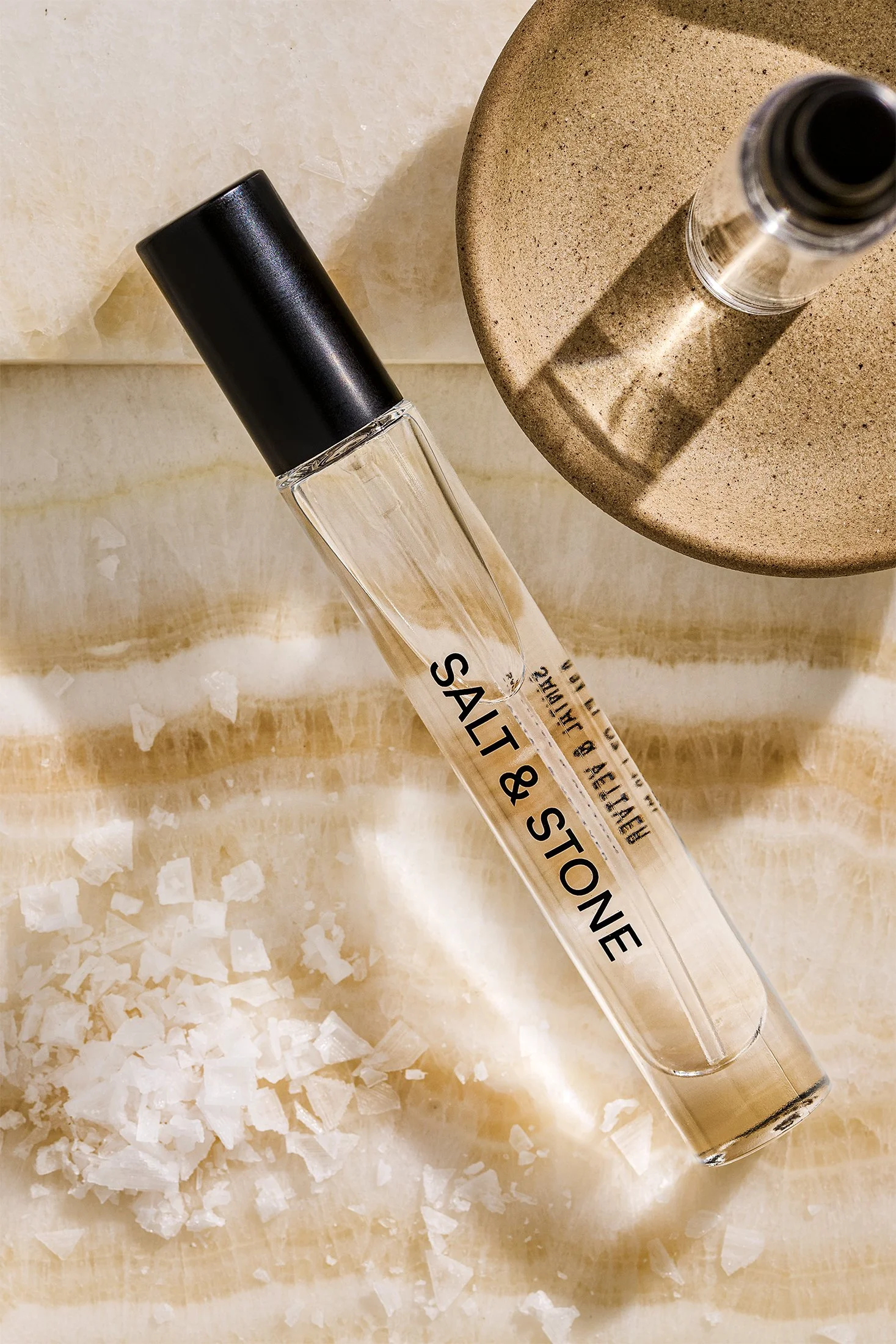 A glass tube labeled 'Salt & Stone' with a black cap, placed on a beige surface near white crystal-like salt flakes, with a round textured beige object in the background.