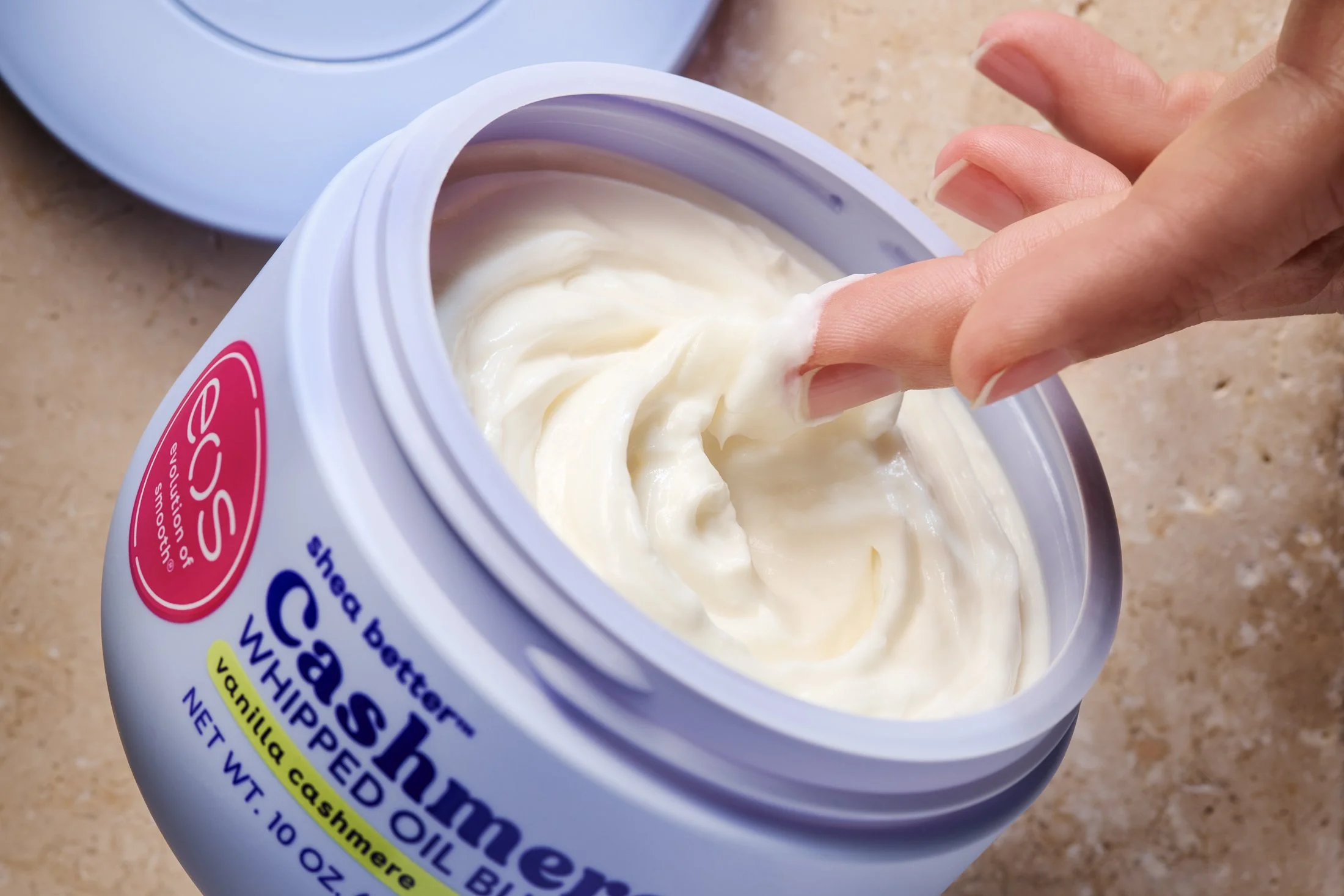 A lotion product in purple container against a pink stone surface with woman's hand dipping into lotion. Product beauty photography by Nutmeg Imageworks.