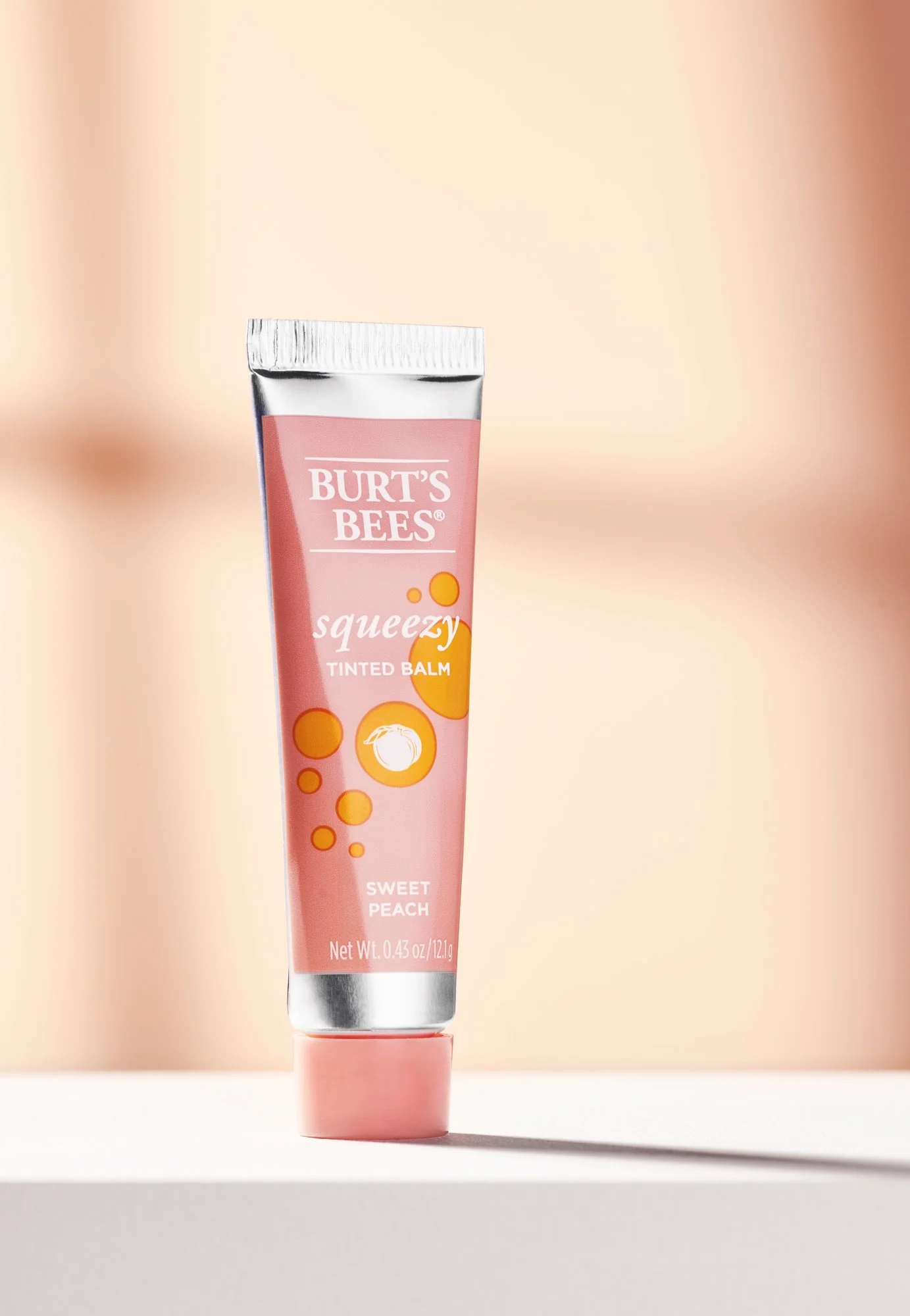 A tube of Burt's Bees tinted lip balm in scent Sweet Peach standing upright on a white shelf against a soft pink background. Beauty product photography by Nutmeg Imageworks.