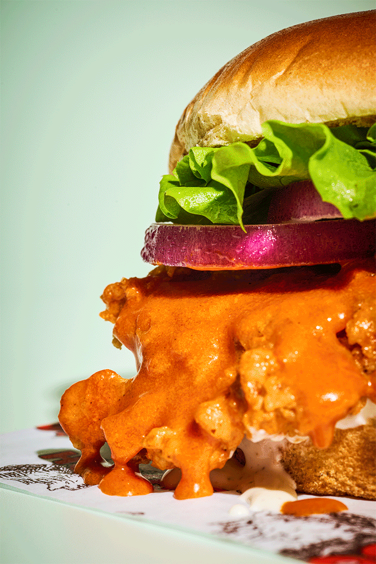Close-up of a burger with lettuce, tomato, and fried chicken breast topped with spicy orange sauce. Food photography gif.