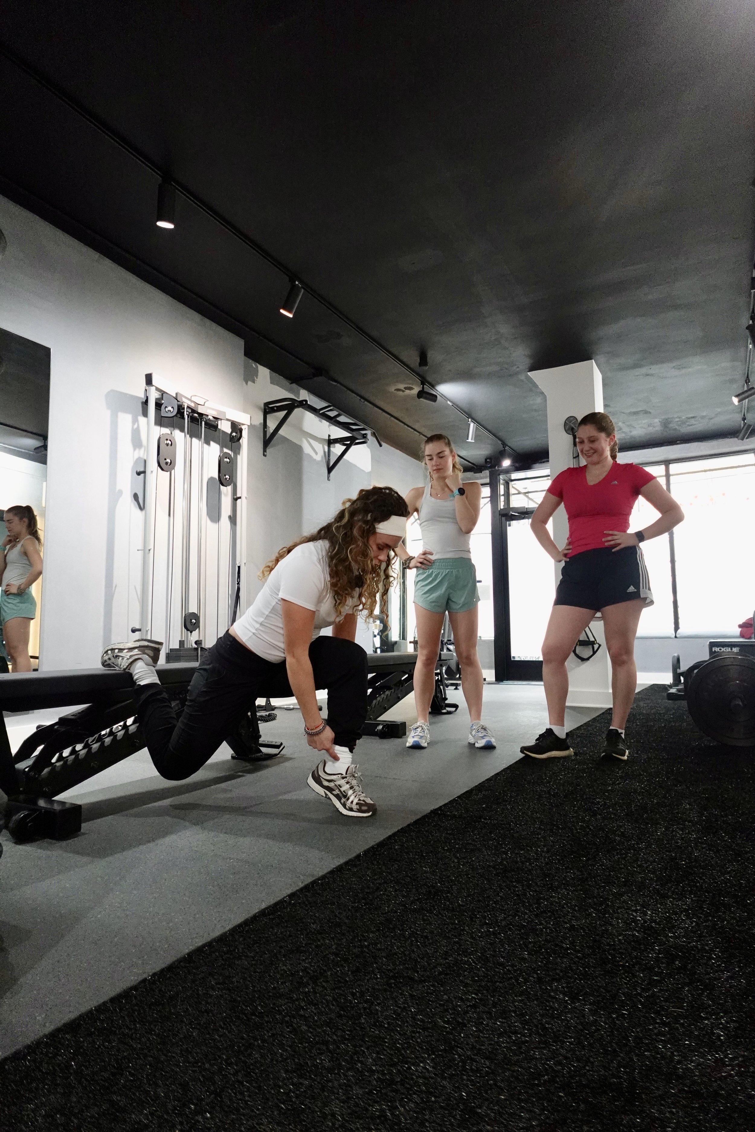 Three women in workout attire in a gym. One woman is adjusting her shoe, while the other two are standing and observe. Gym equipment with weights and mirrors are visible in the background.