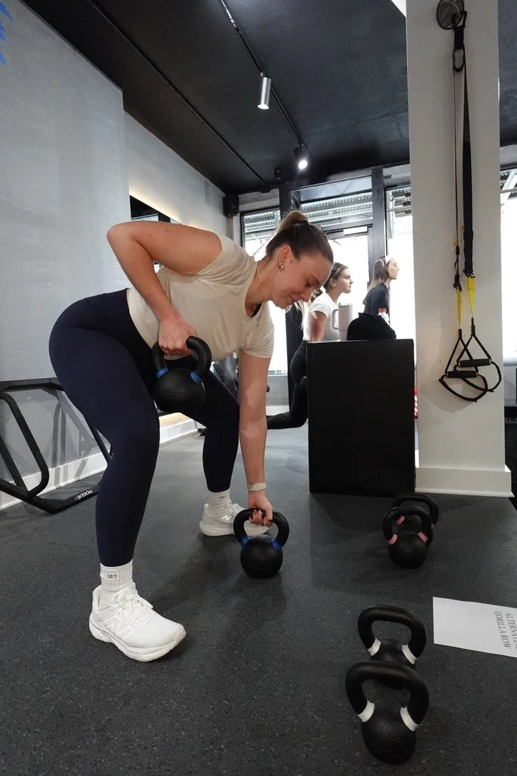 A woman in workout attire is performing a kettlebell exercise in a gym, with other women working out in the background.