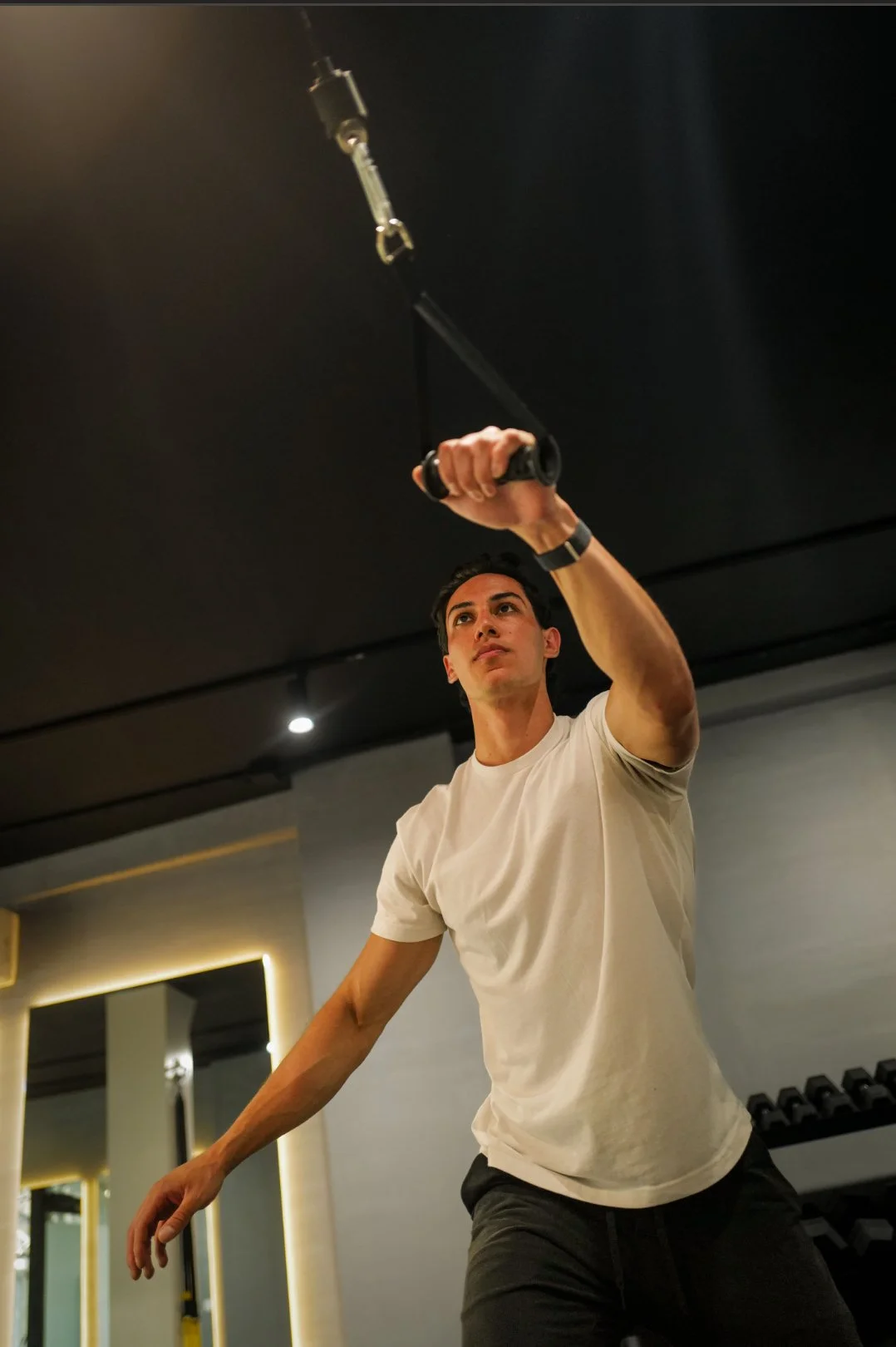 Young man in a white shirt working out at the gym, using a cable machine for a triceps extension exercise.
