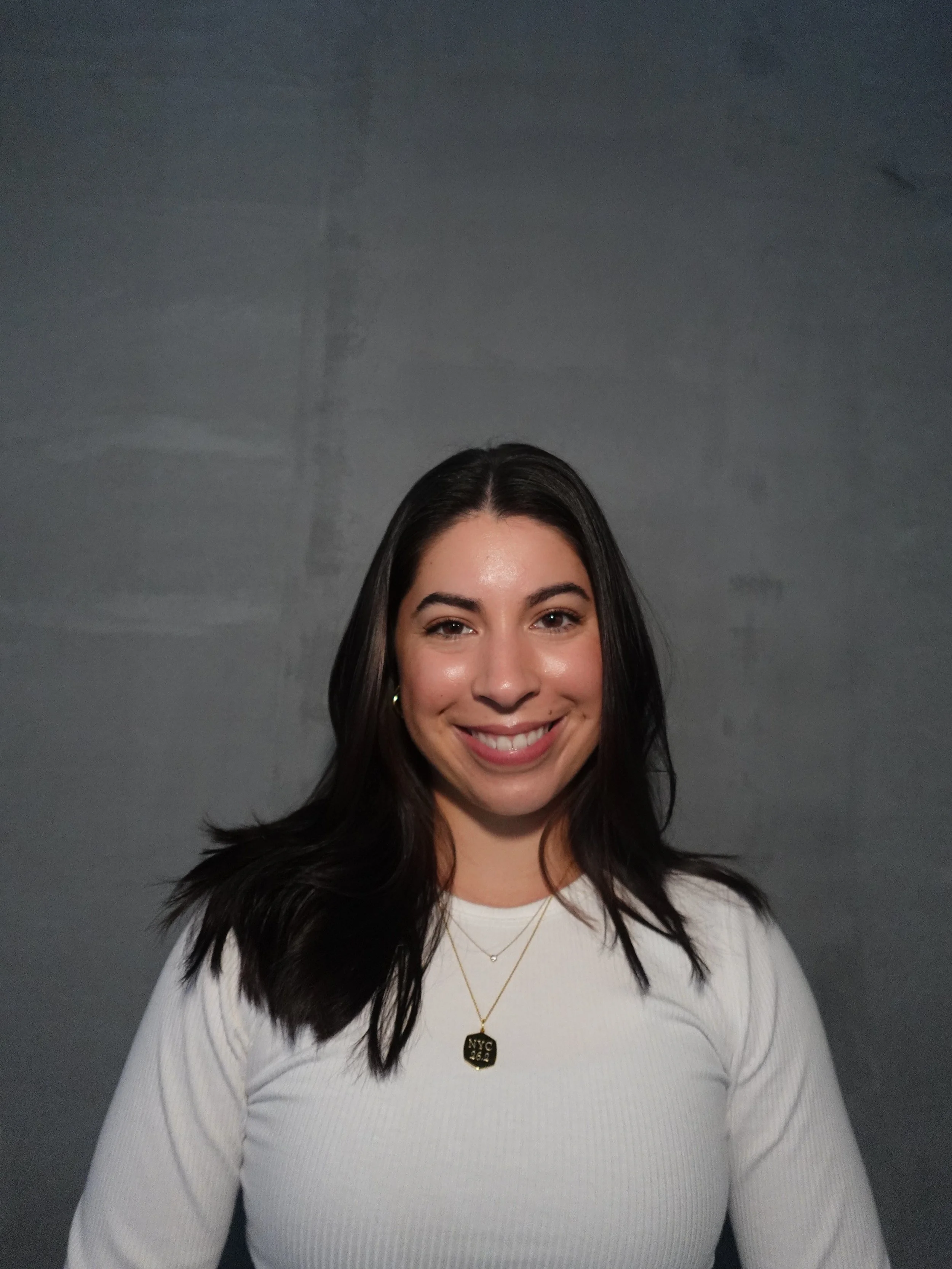 A woman with dark hair smiling at the camera, wearing a white top and layered necklaces, standing against a plain gray background.