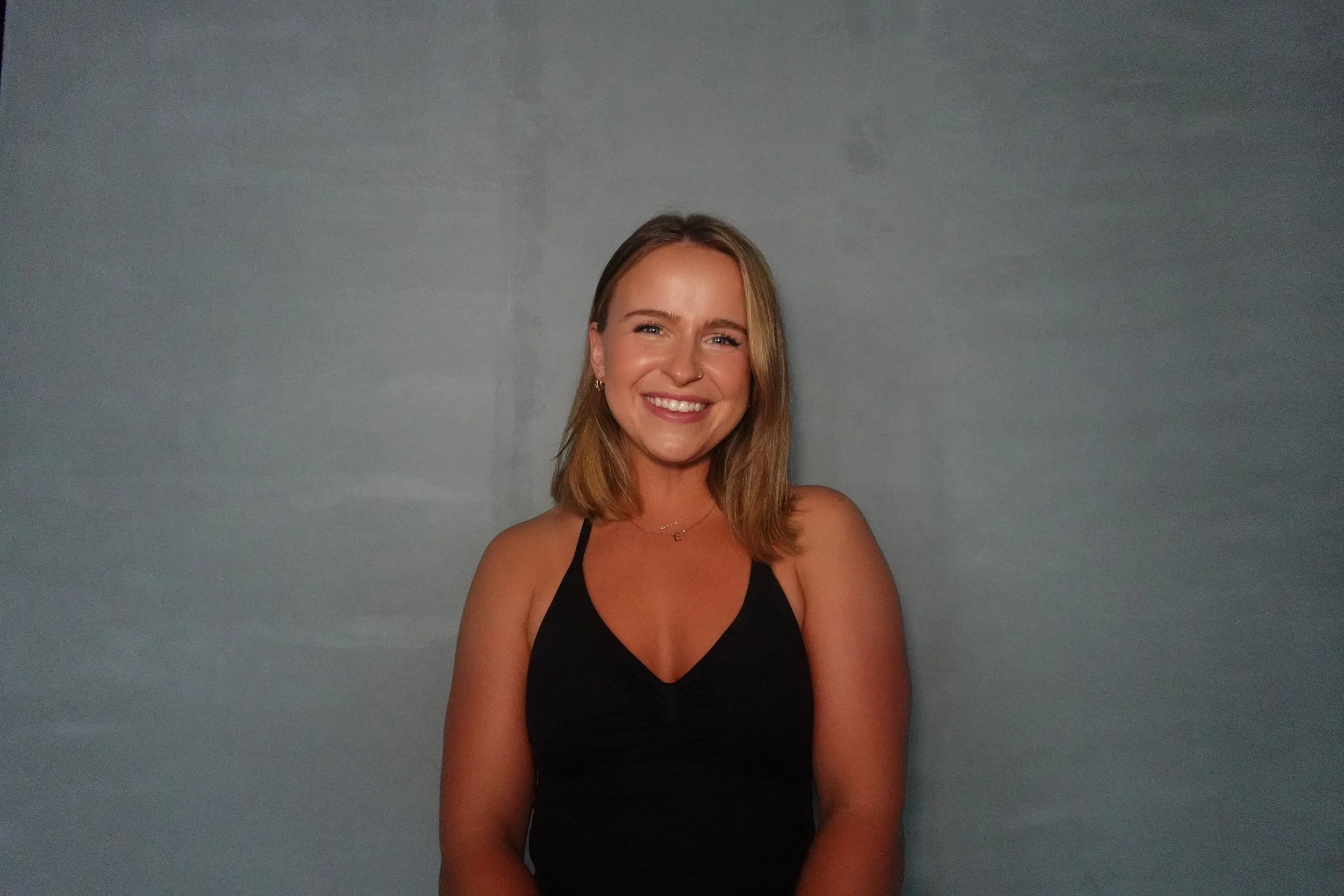 A young woman with shoulder-length blonde hair, smiling, wearing a black sleeveless top, standing against a plain gray wall.