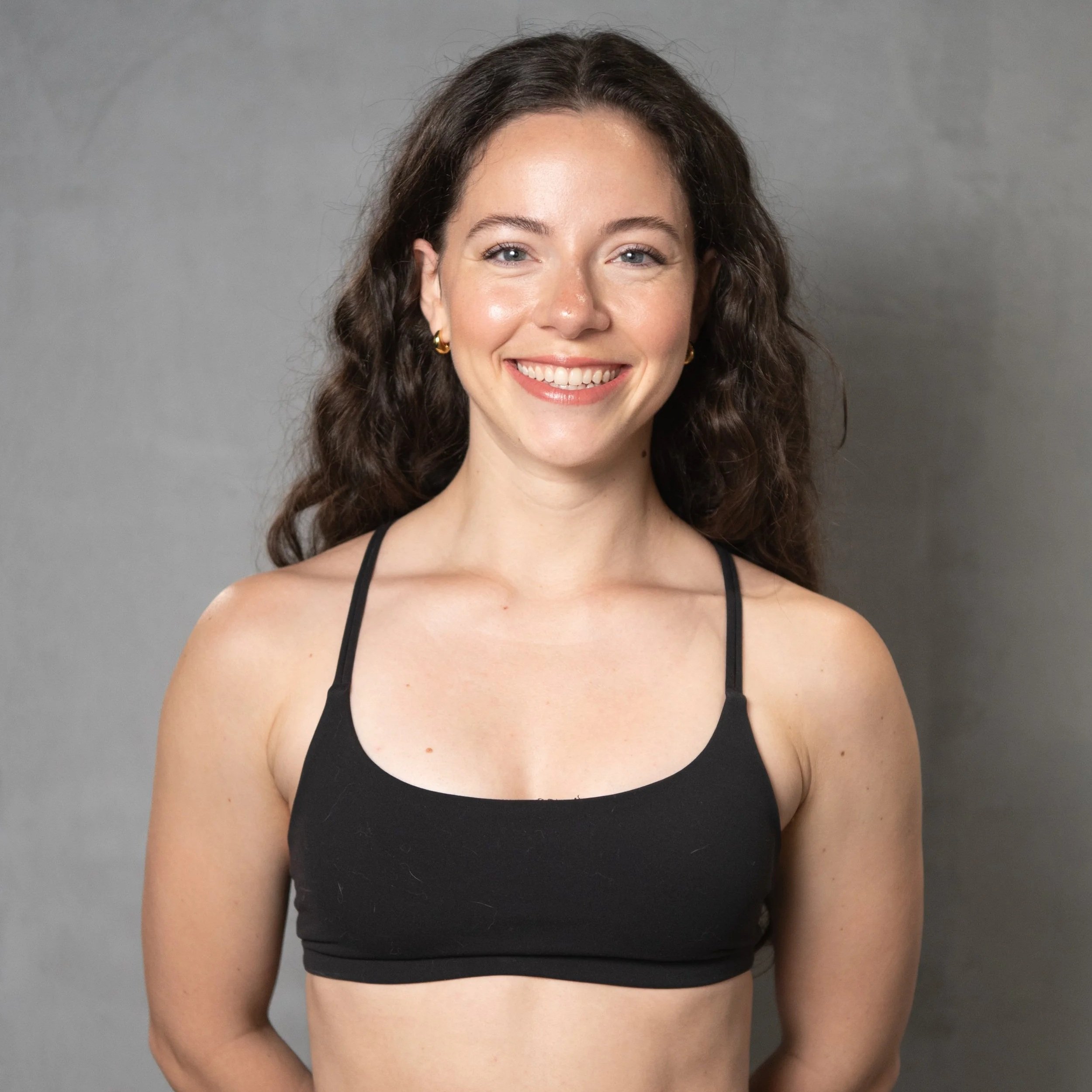 A woman with long, curly brown hair and light skin, smiling, wearing a black sports bra, standing against a gray background.