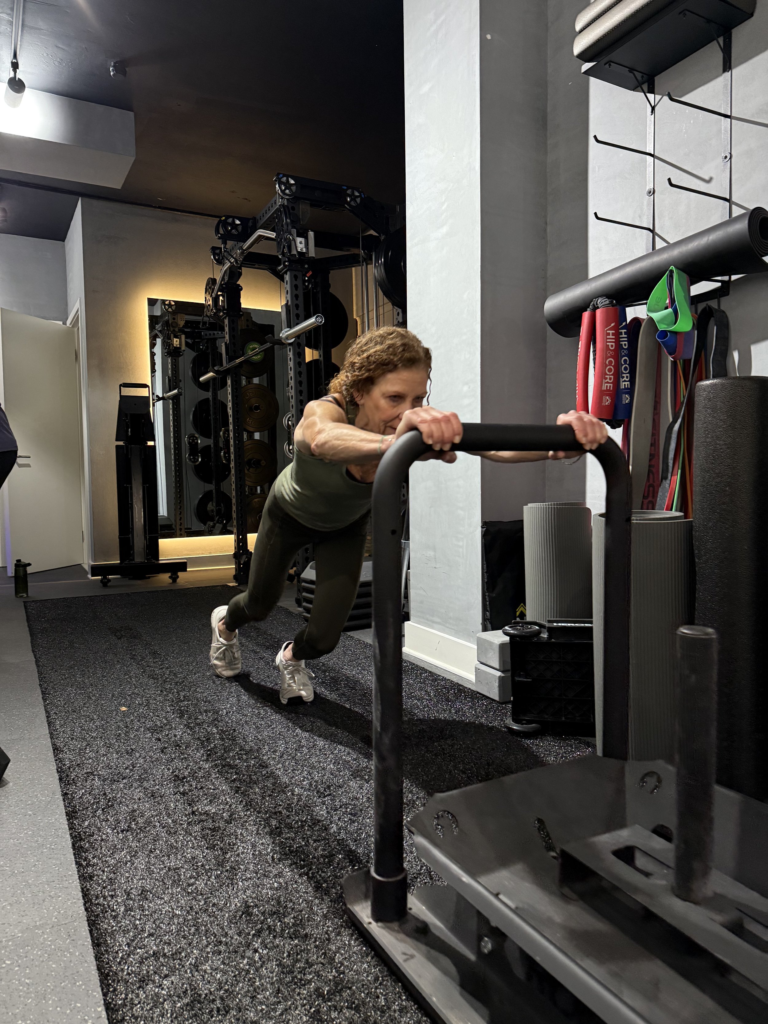 An elderly woman with curly hair wearing a green tank top and black leggings doing a push-up exercise on a padded floor in a gym, with gym equipment and weights in the background.