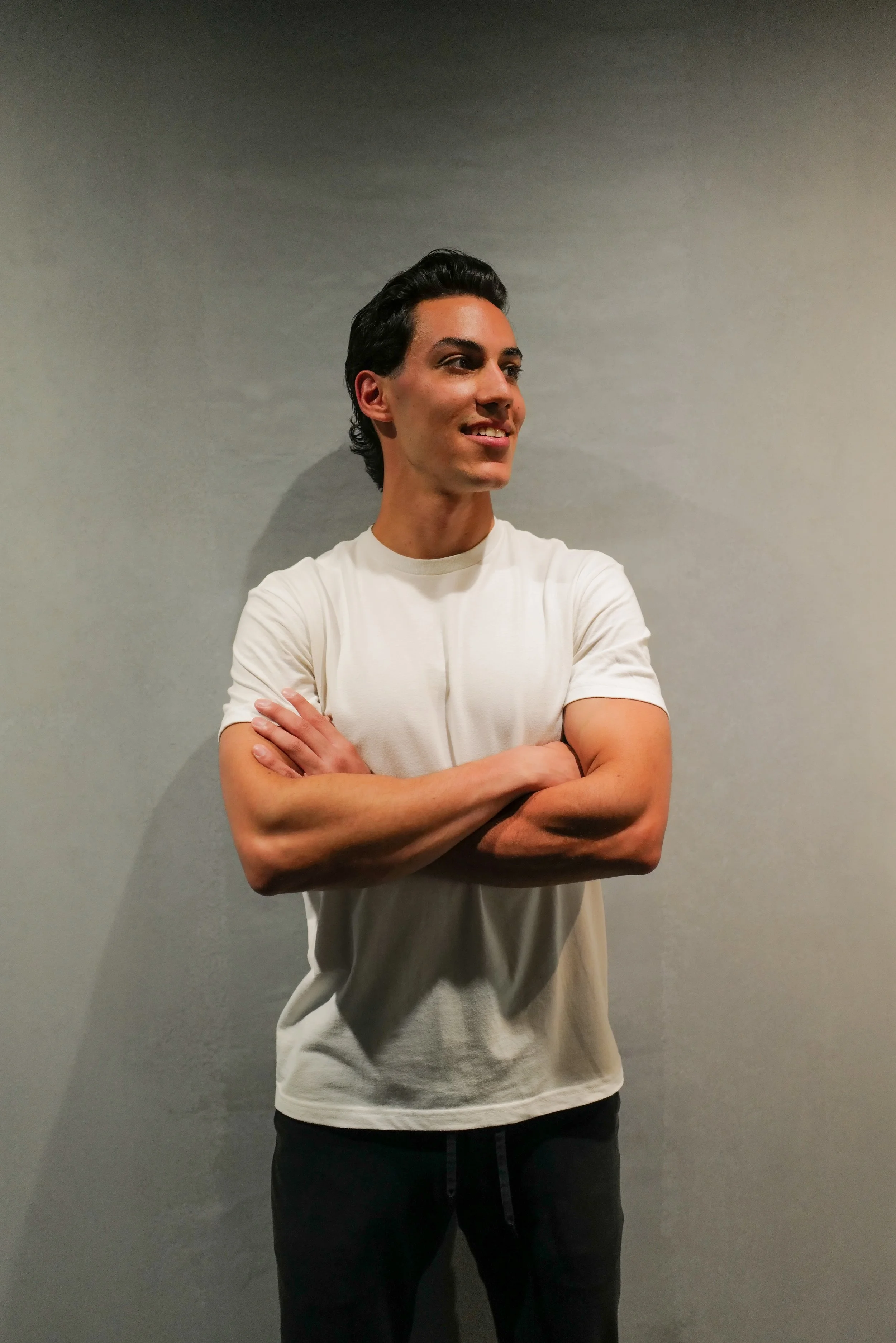 A young man with dark hair smiling and crossing his arms, wearing a white t-shirt and black pants, standing against a plain light gray wall.