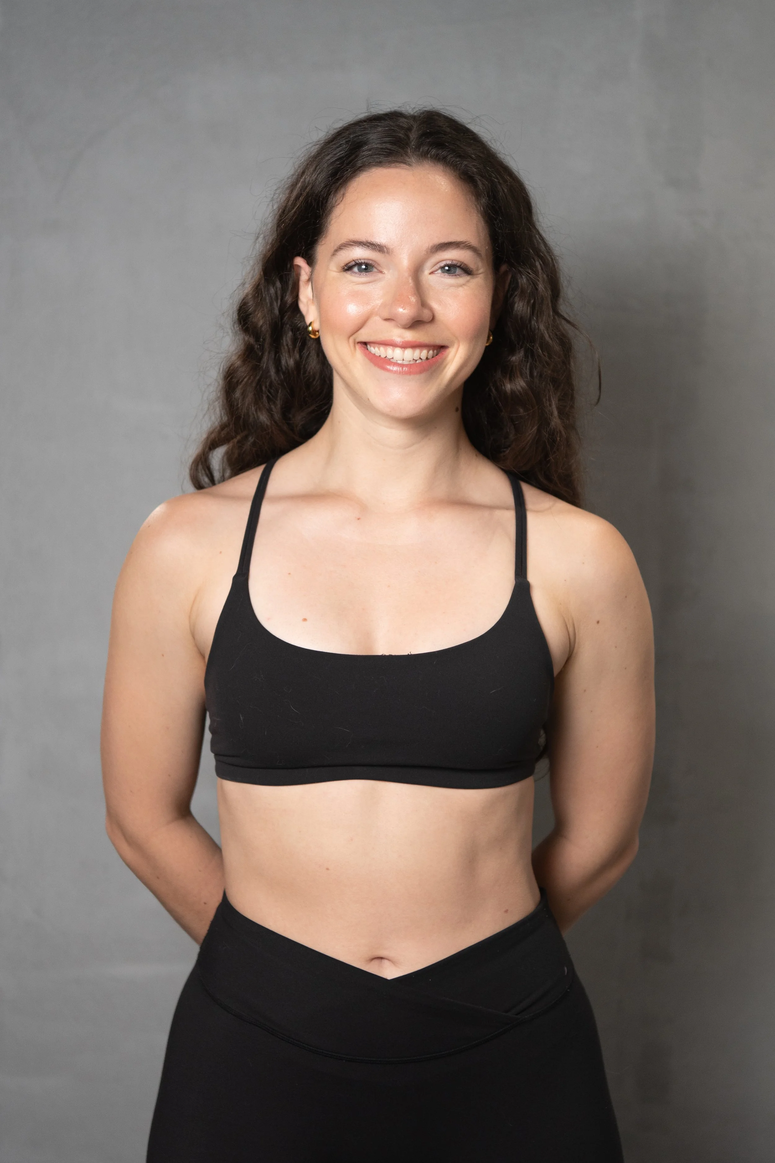 A woman smiling, wearing a black sports bra and black leggings, standing against a plain grey background.