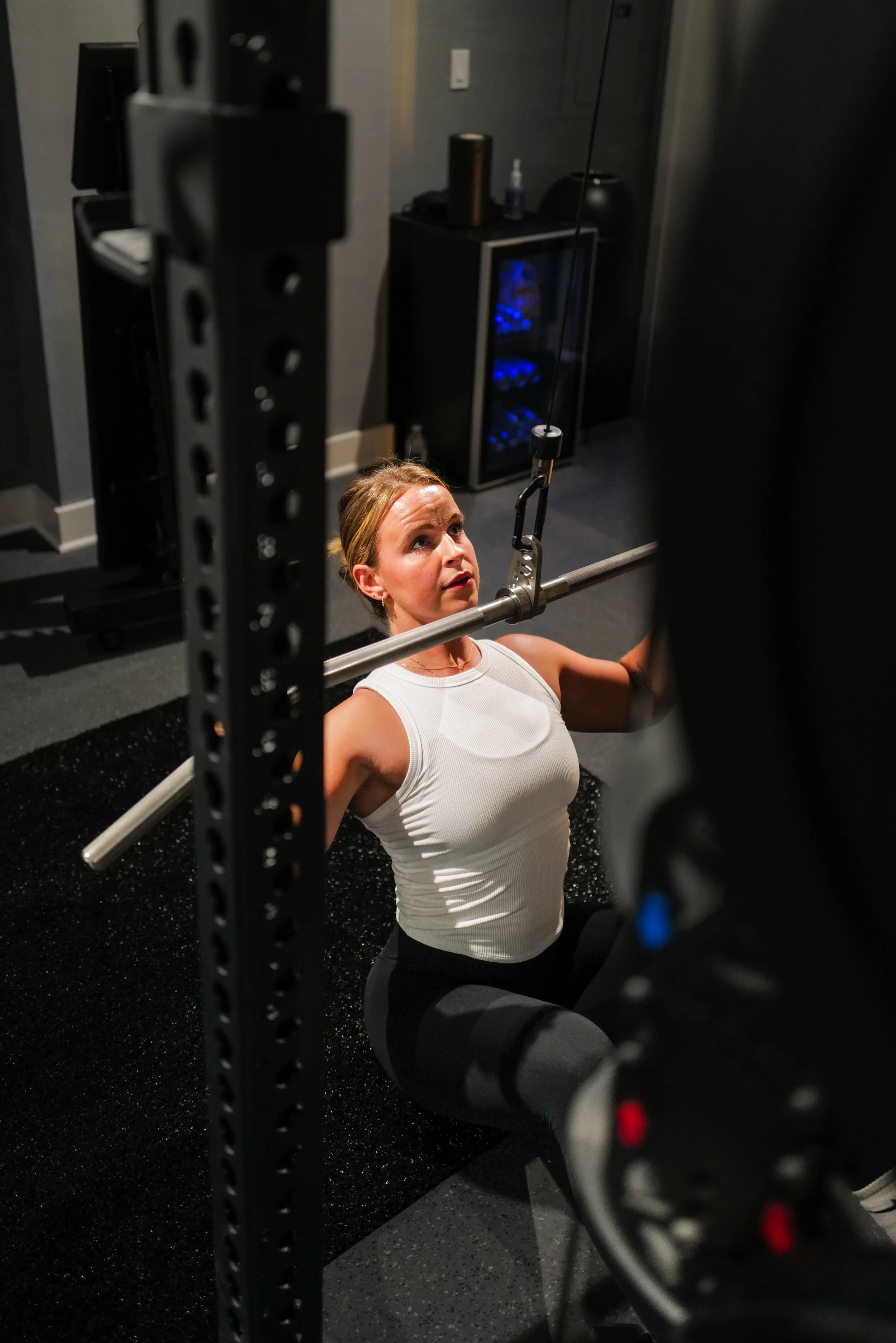 A woman in a white sleeveless top and black leggings is exercising in a gym, lifting a barbell while kneeling on a black mat.