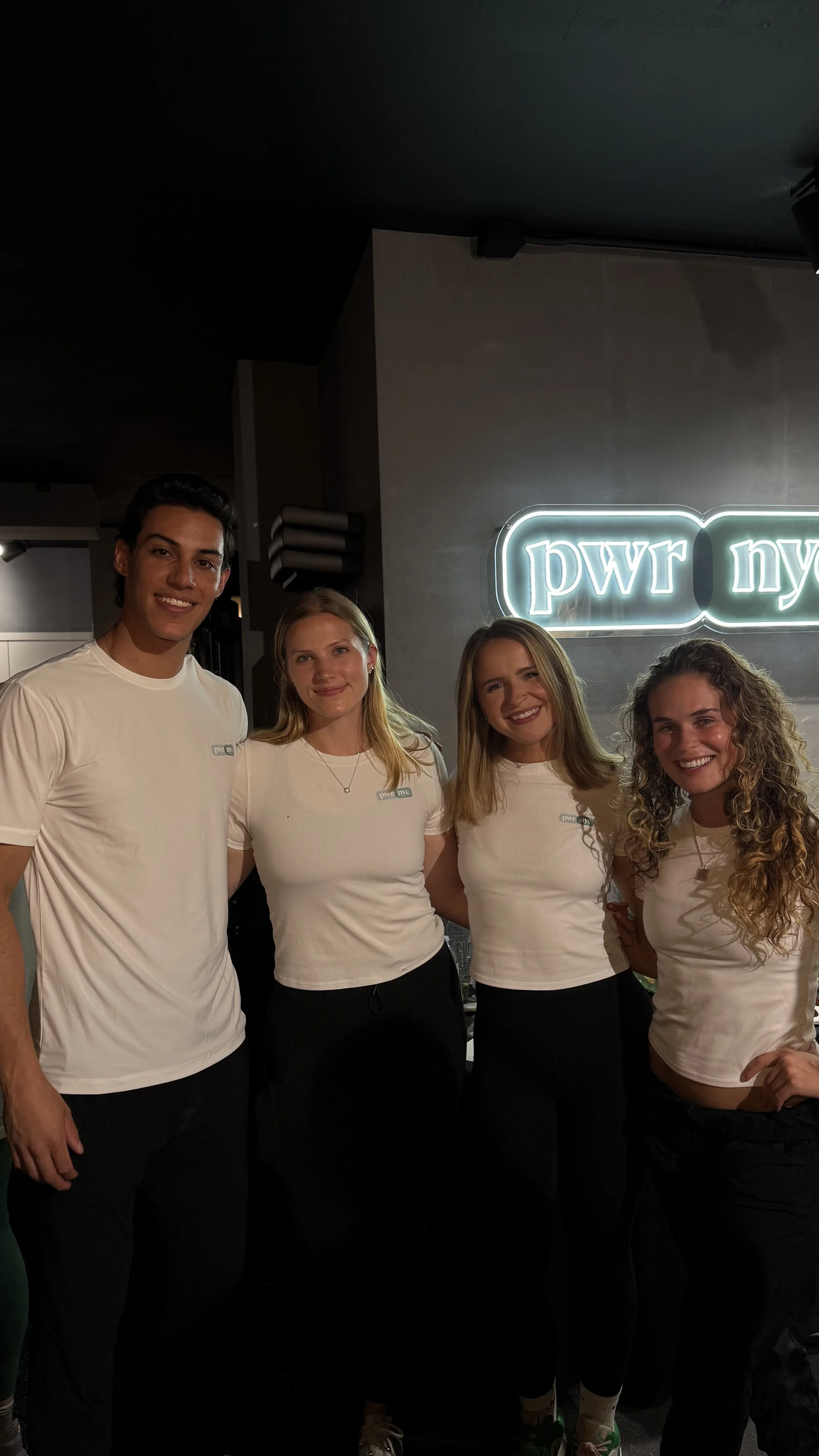 Four young adults, two men and two women, standing close together in front of a wall with a glowing neon sign that says "pwr nyc." They are all smiling and wearing white t-shirts.