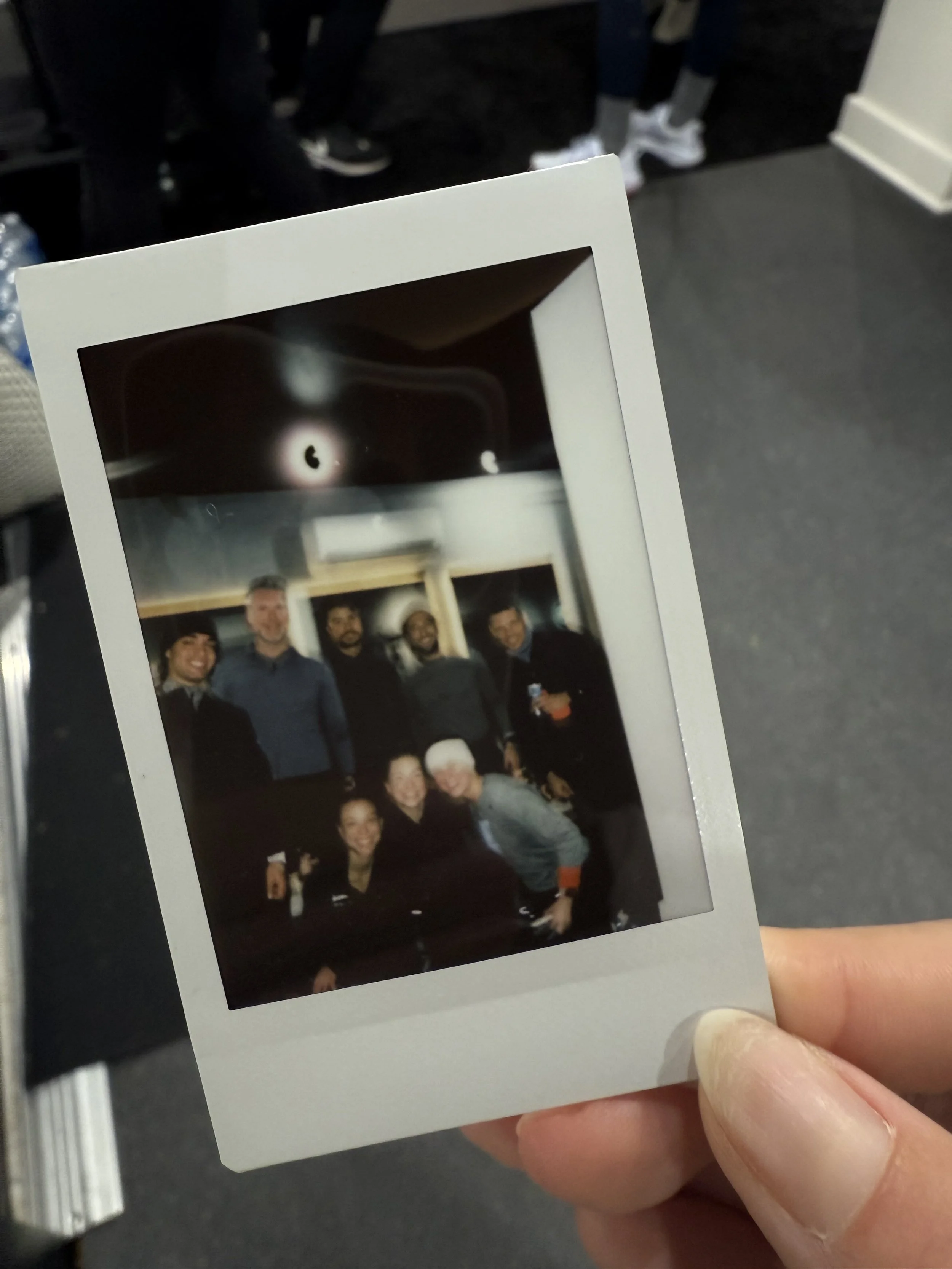 Casual group photo of seven people smiling, gathered indoors in a room with black walls and shelving, some holding drinks.