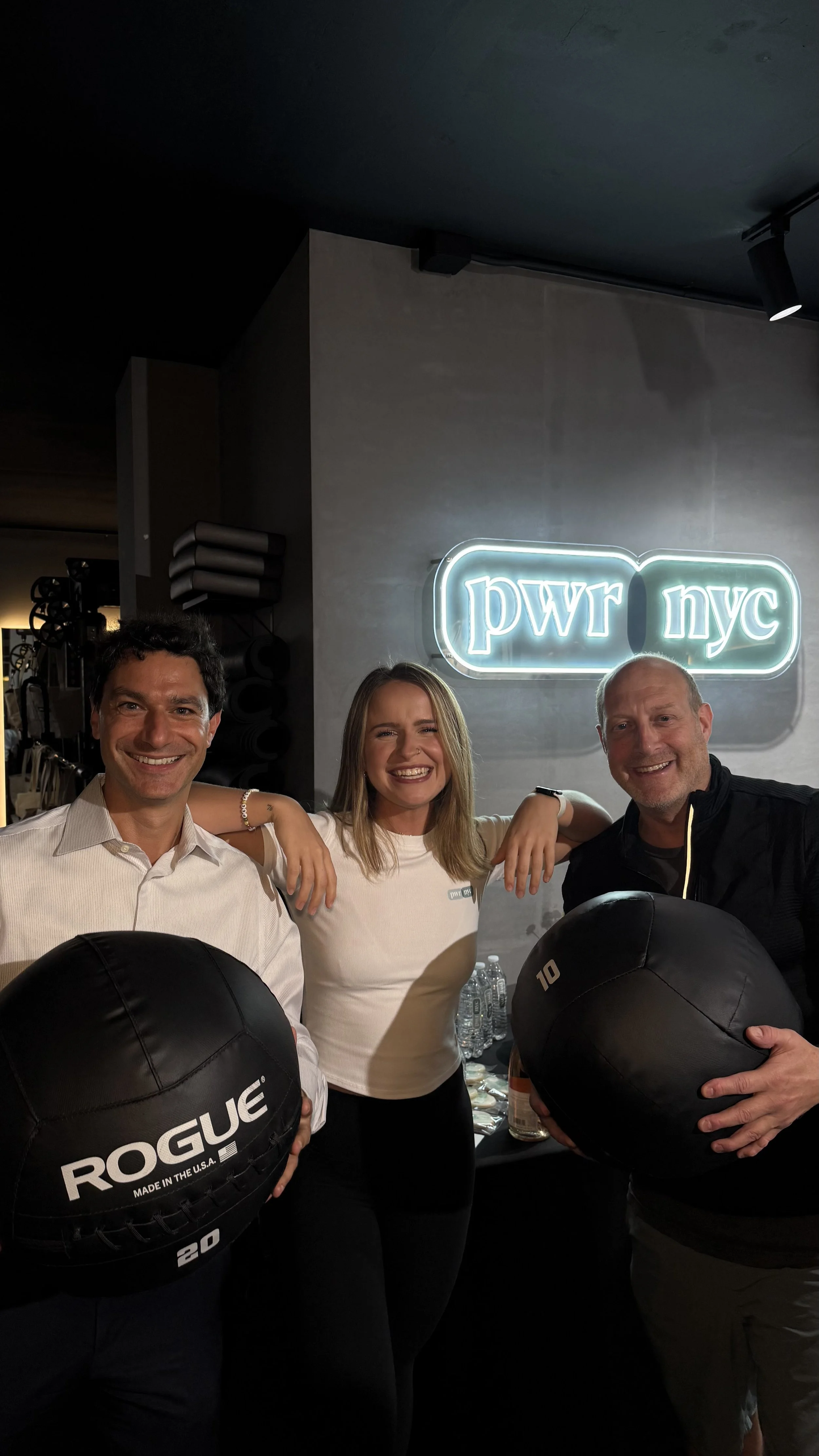Three people smiling and posing with medicine balls in front of a neon sign that reads 'pwr nyc,' in a fitness or gym setting.