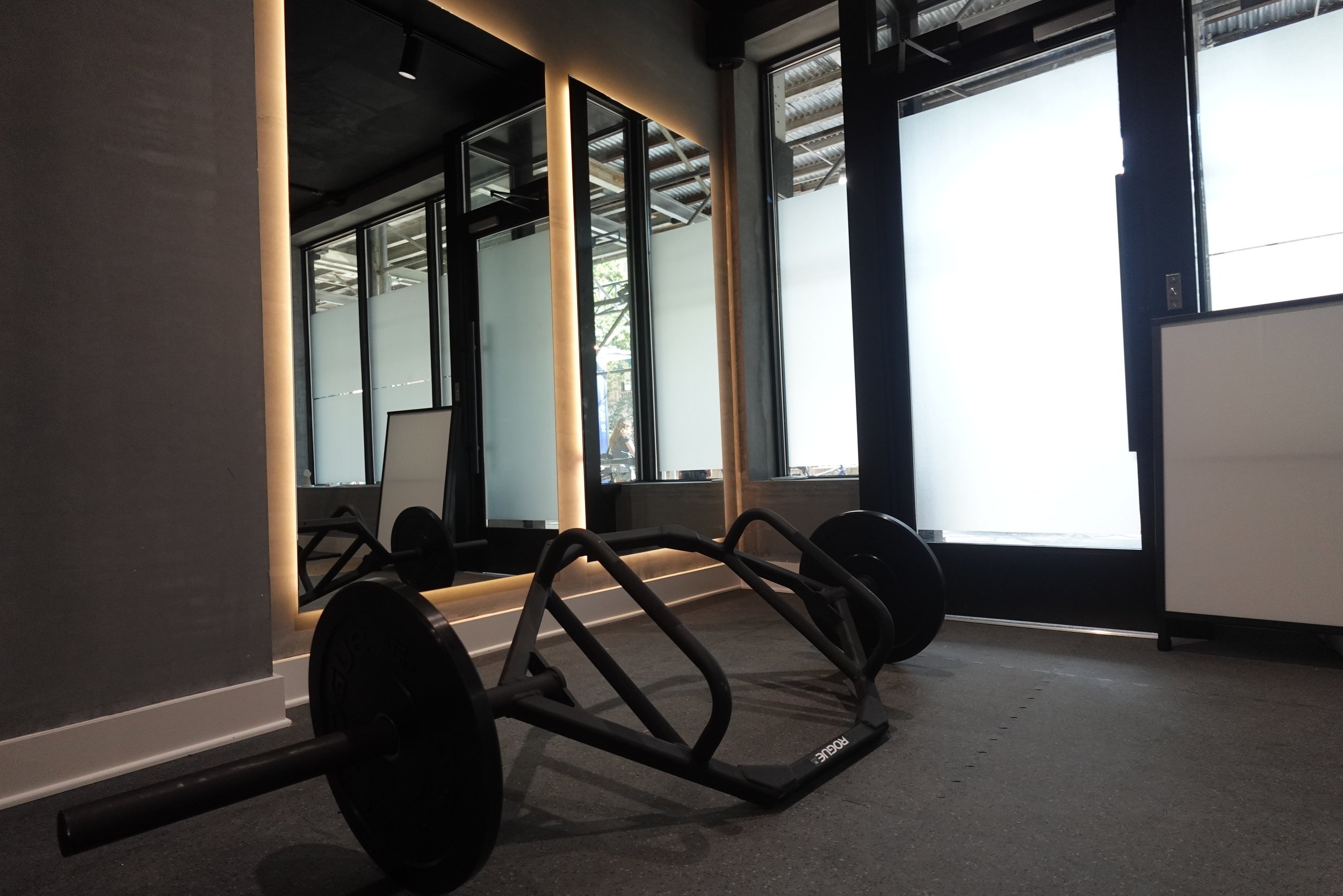 Empty gym corner with black weightlifting barbell and squat rack on a gray carpeted floor, large window with frosted glass, and a large mirror with warm lighting around it.